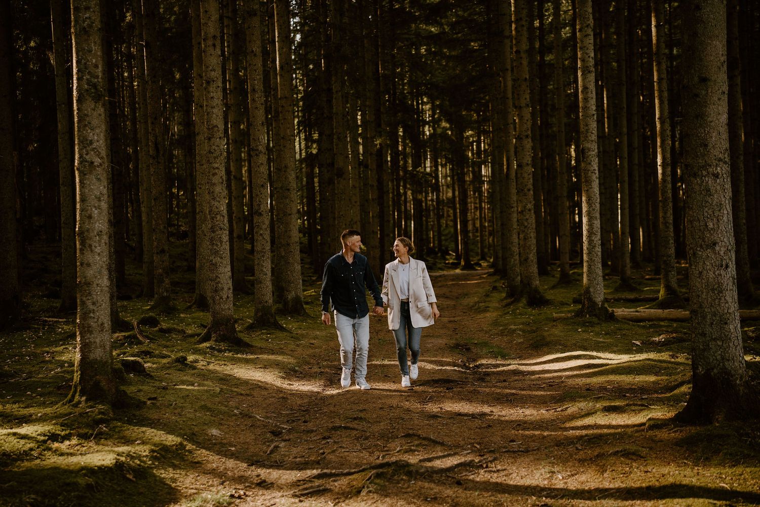 Two people walk hand in hand down a sunlit forest path surrounded by tall pine trees on a peaceful afternoon.