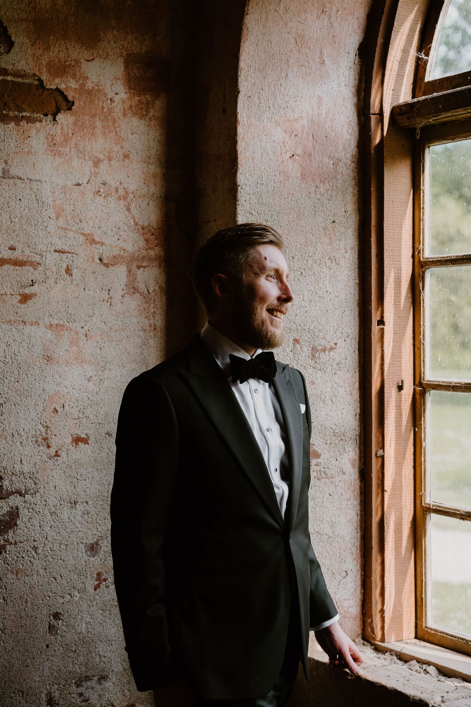 A person in formal attire stands in a rustic room with sunlit windows and weathered brick walls.