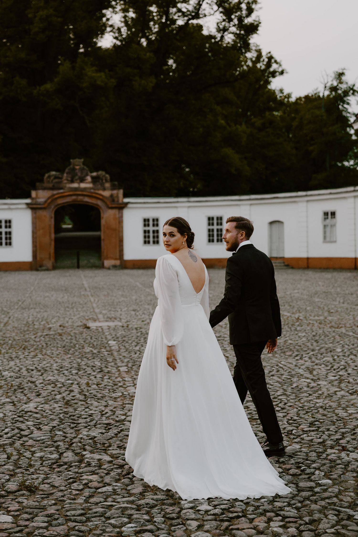 Two figures in wedding attire walk together on a cobblestone courtyard against a white building facade.