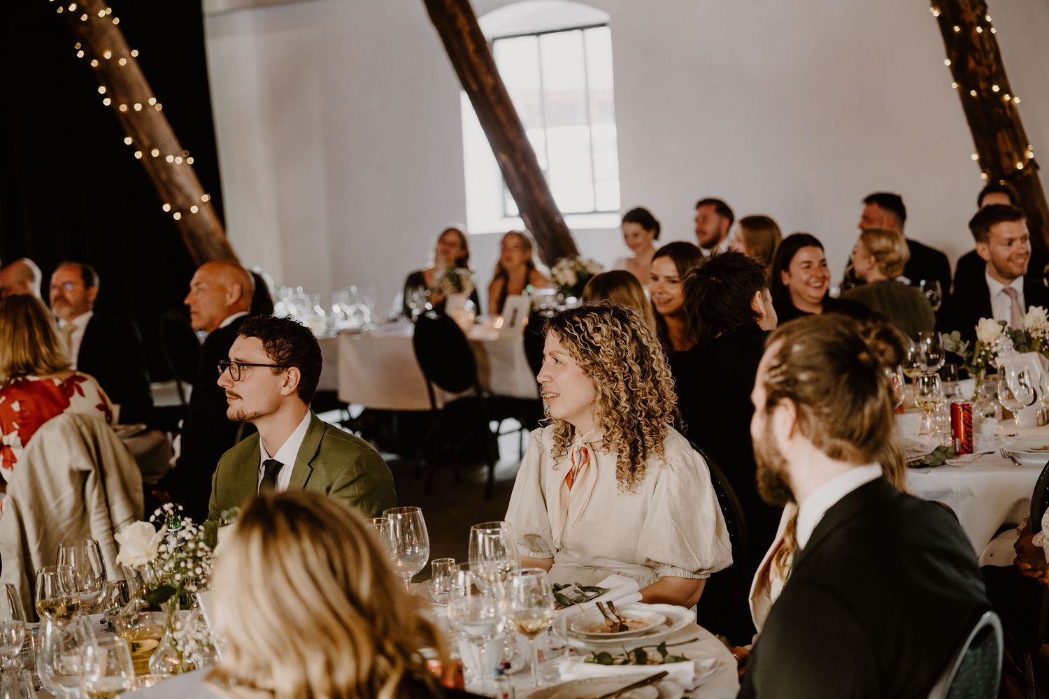 Wedding guests seated at elegant dining tables with string lights glowing overhead during an indoor reception celebration.