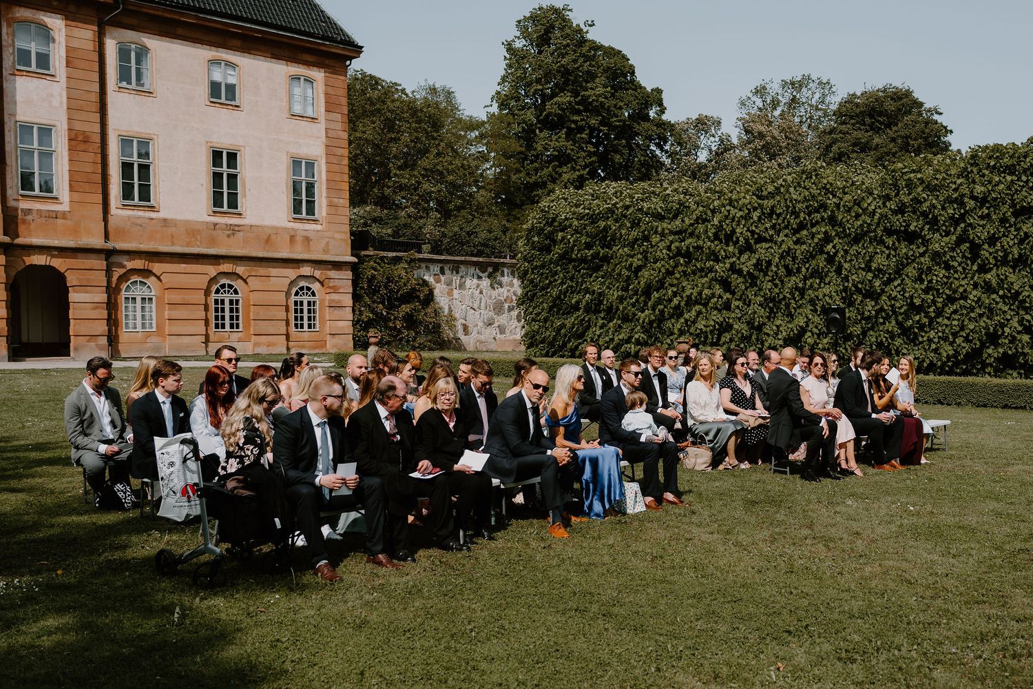 Guests seated on white chairs for an outdoor wedding ceremony on a lawn in front of a historic pink mansion.