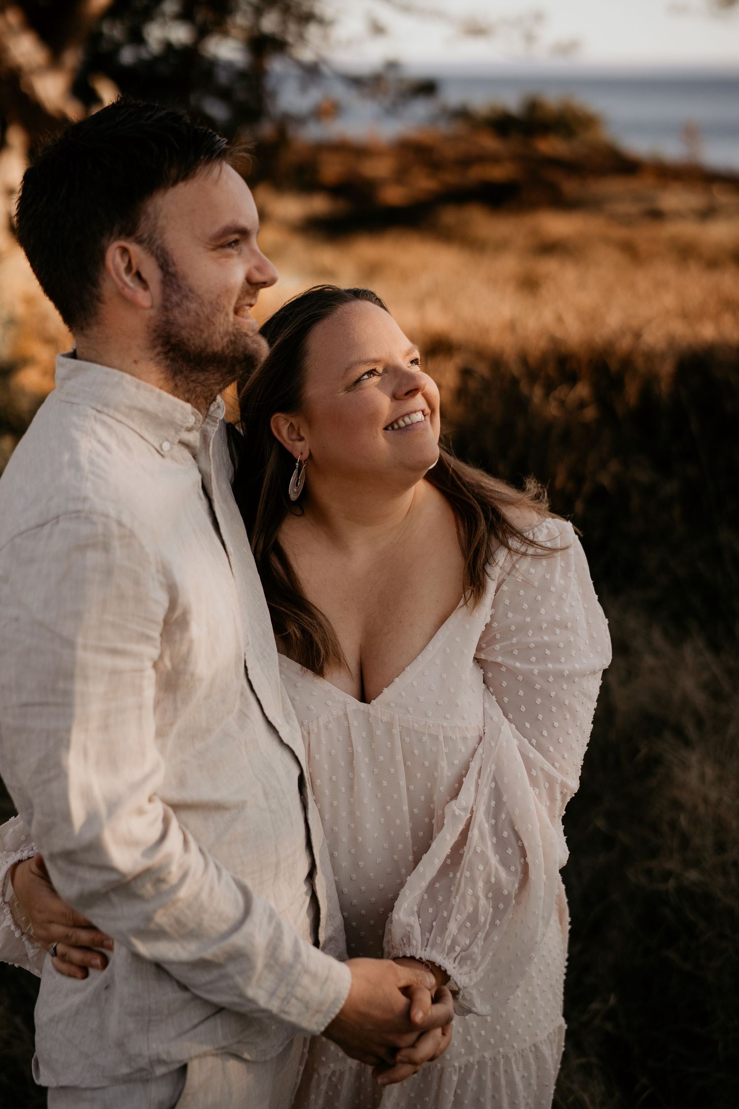 A couple in white outfits sharing a tender moment at sunset along a scenic coastal landscape.