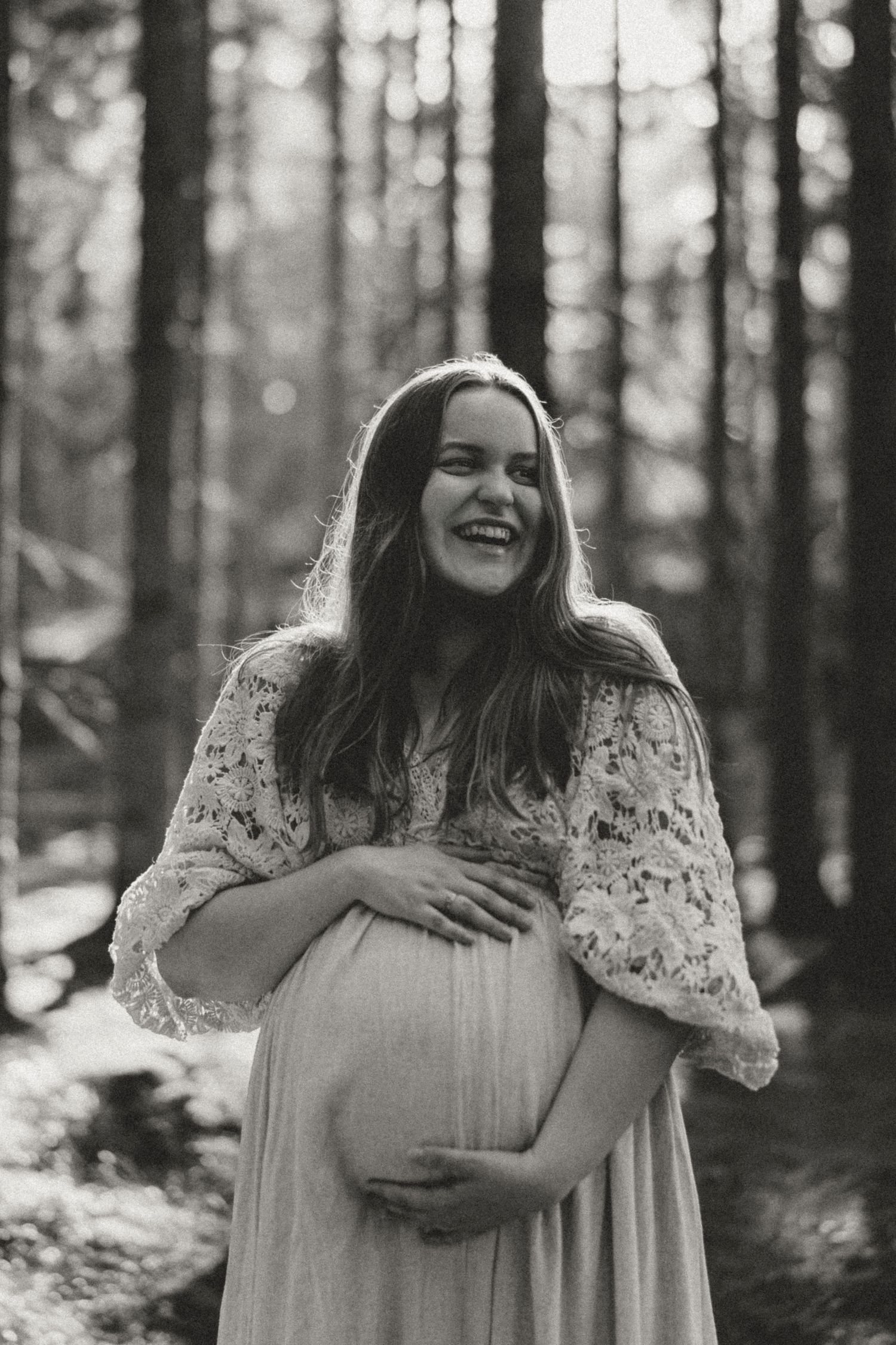 Black and white photo of expecting mother in lace dress cradling baby bump while laughing in sunlit forest.