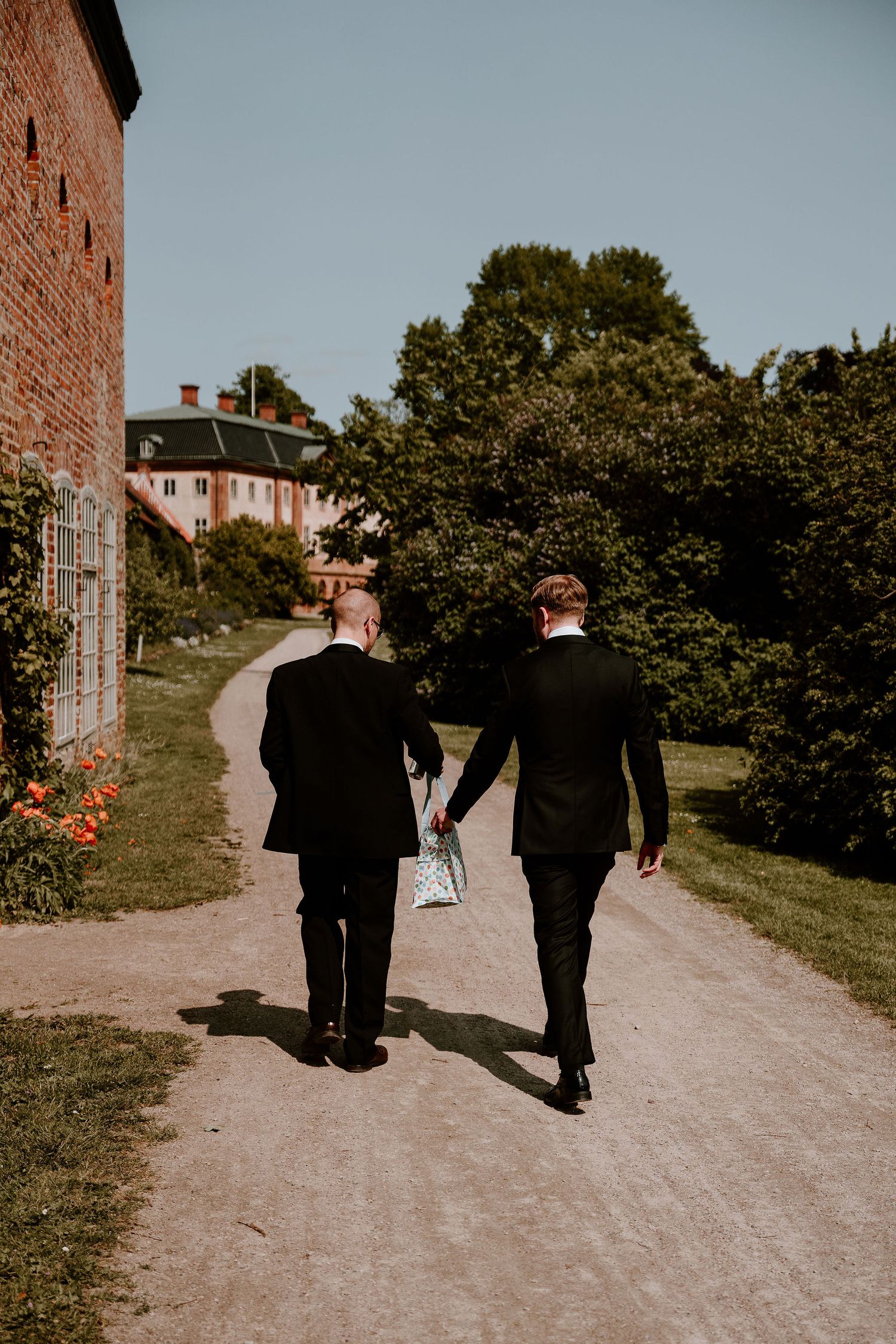 Two people walking down a dirt path on a sunny day with trees and buildings in the background.