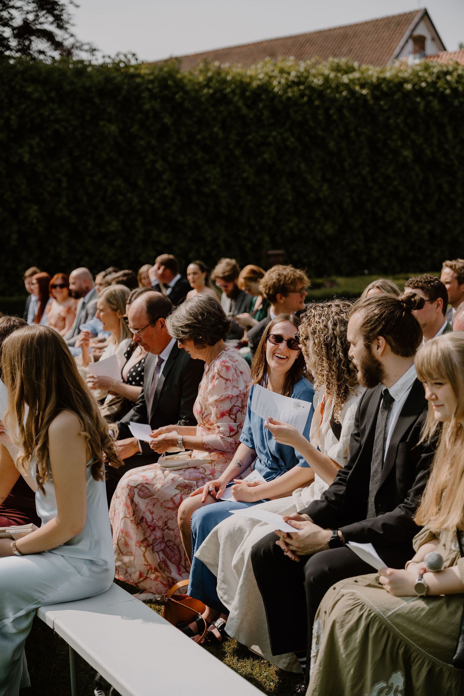 Wedding guests seated outdoors on white chairs smile and chat before the ceremony on a sunny afternoon.