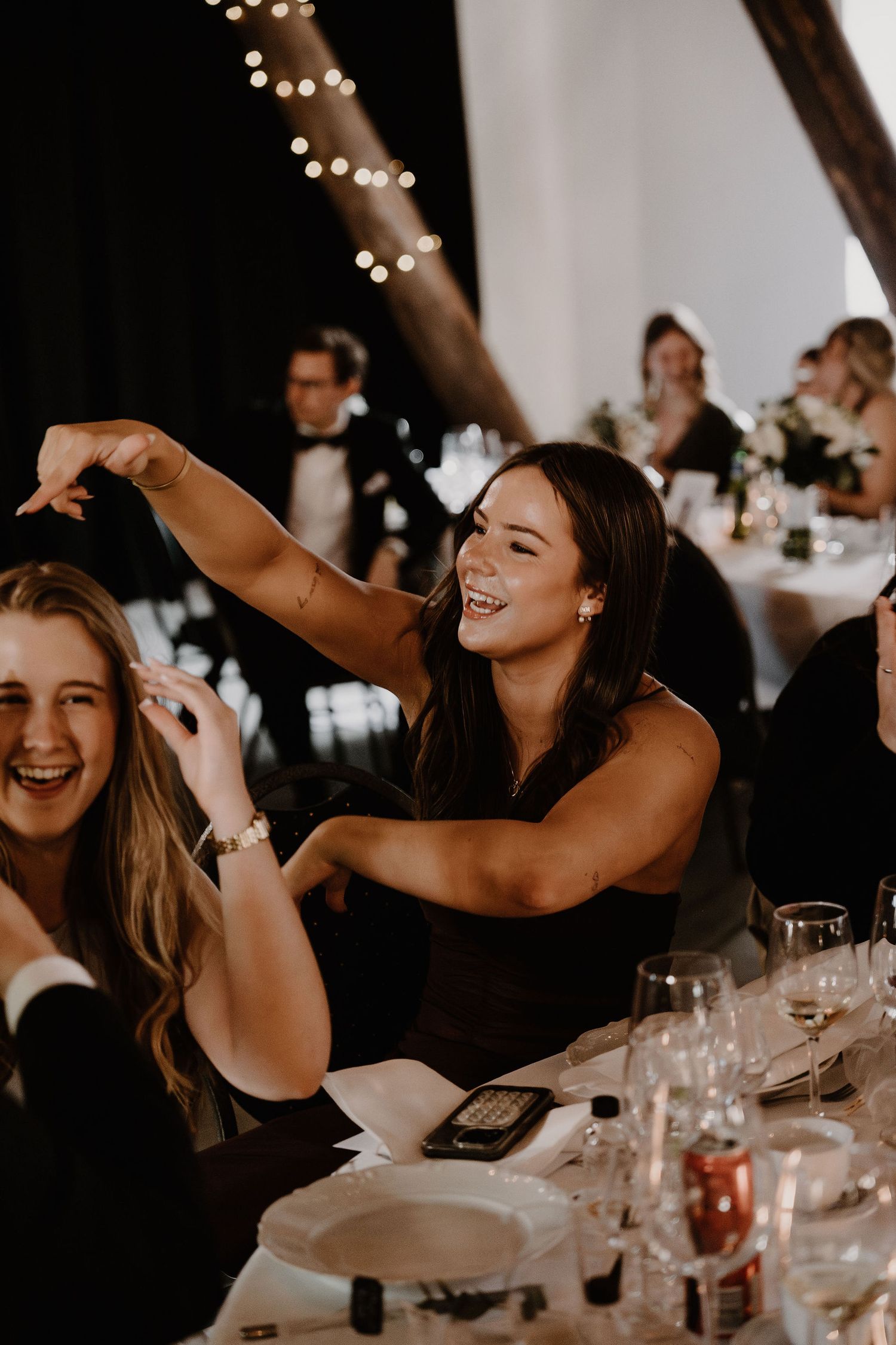 Wedding guests laughing and having fun at an elegantly decorated reception dinner with string lights overhead.