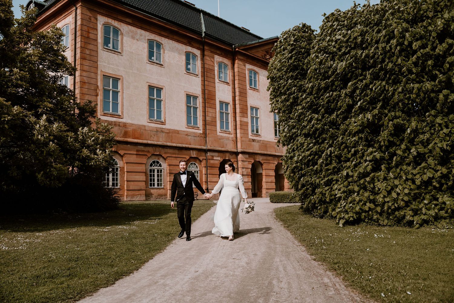 Elegant couple walking on gravel path outside historic brick mansion with tall green trees in summer.