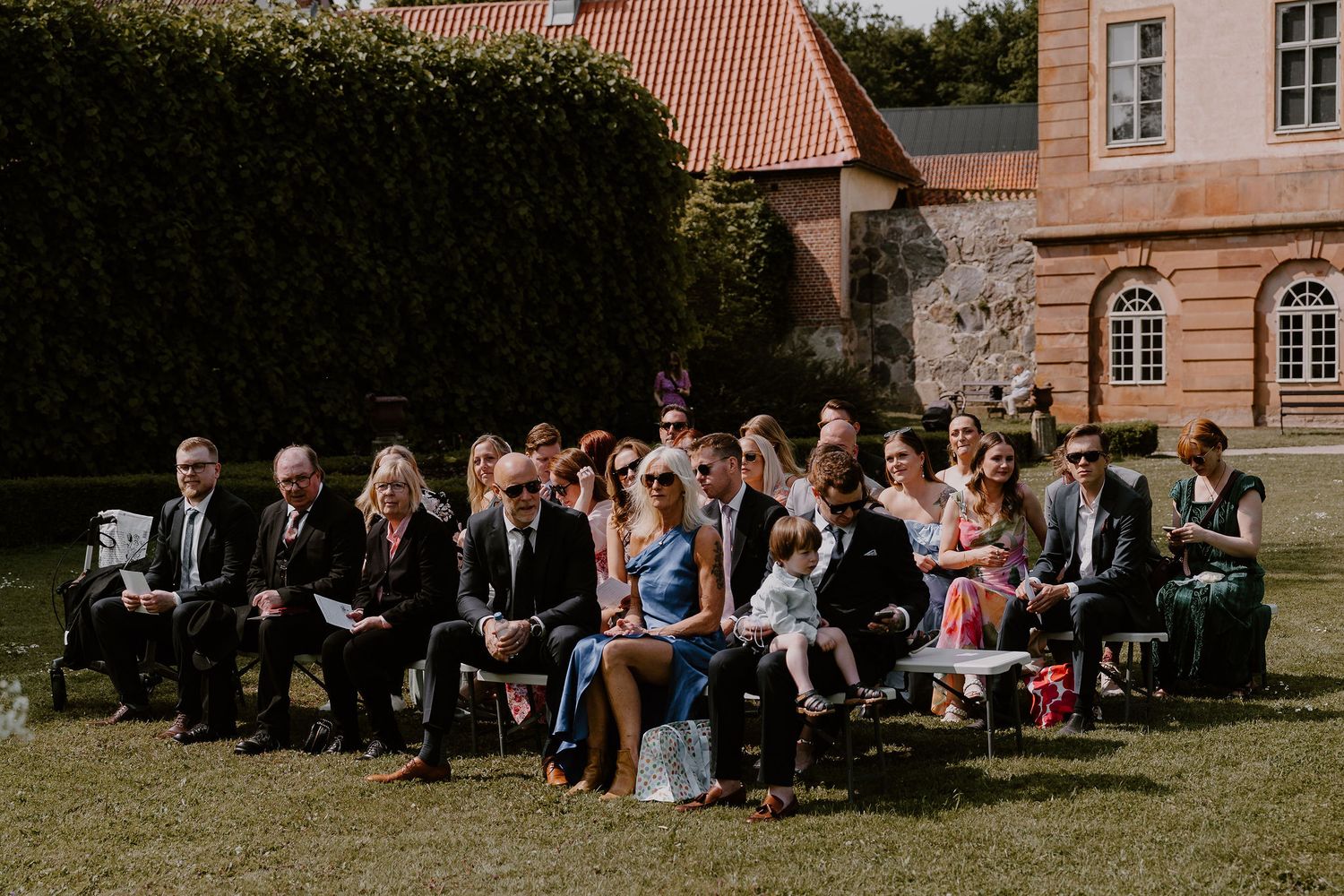 Outdoor wedding ceremony guests seated on folding chairs in a garden setting with historic brick building in background.