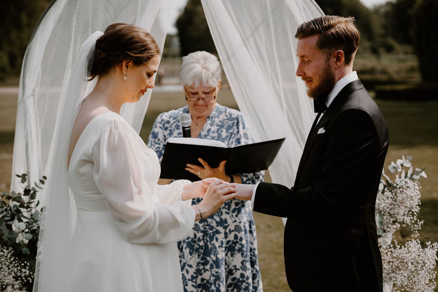 A couple exchanges rings during an outdoor wedding ceremony beneath a white fabric arch.