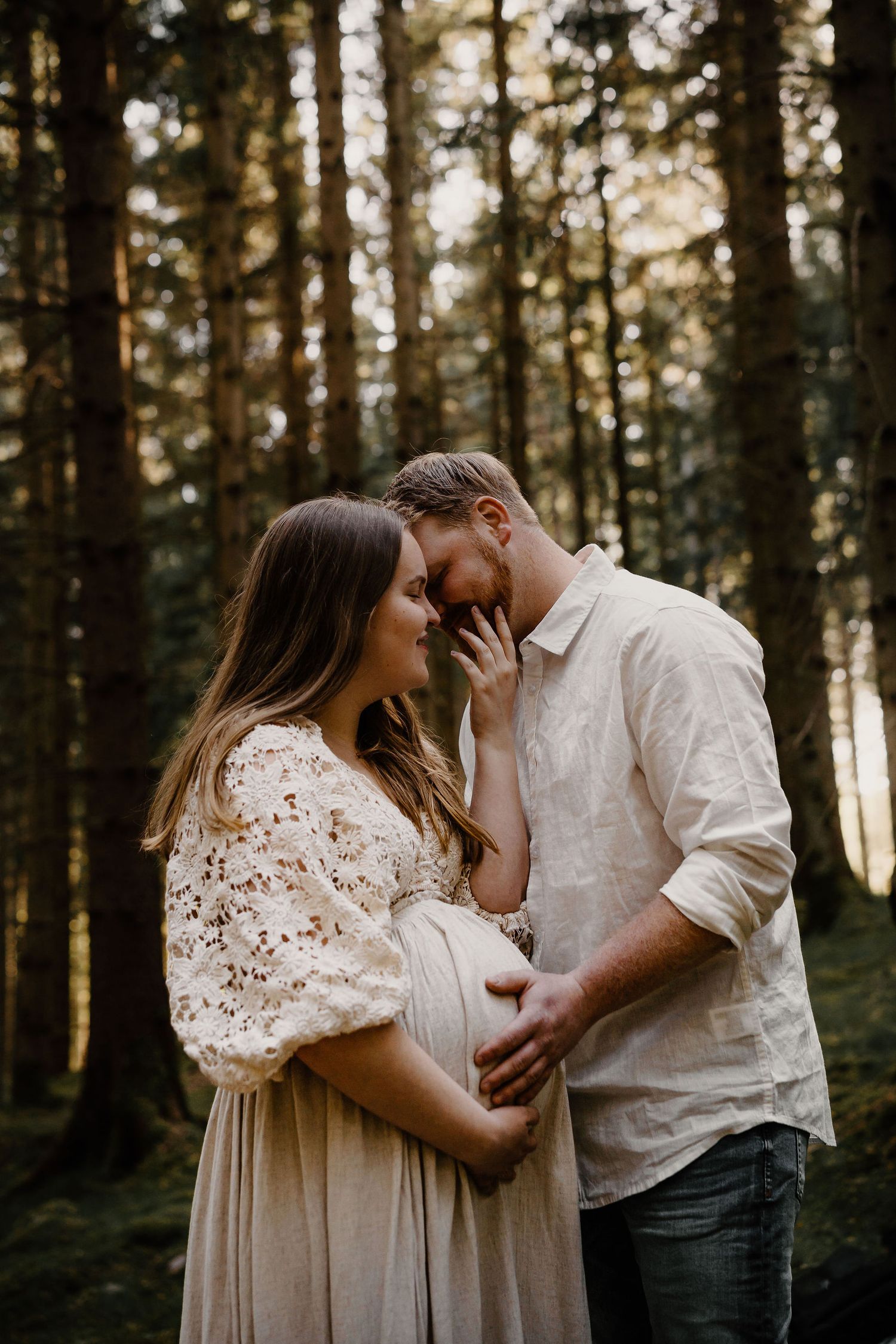A romantic maternity photo in a moody forest setting shows an intimate moment between a couple in neutral-toned clothing.
