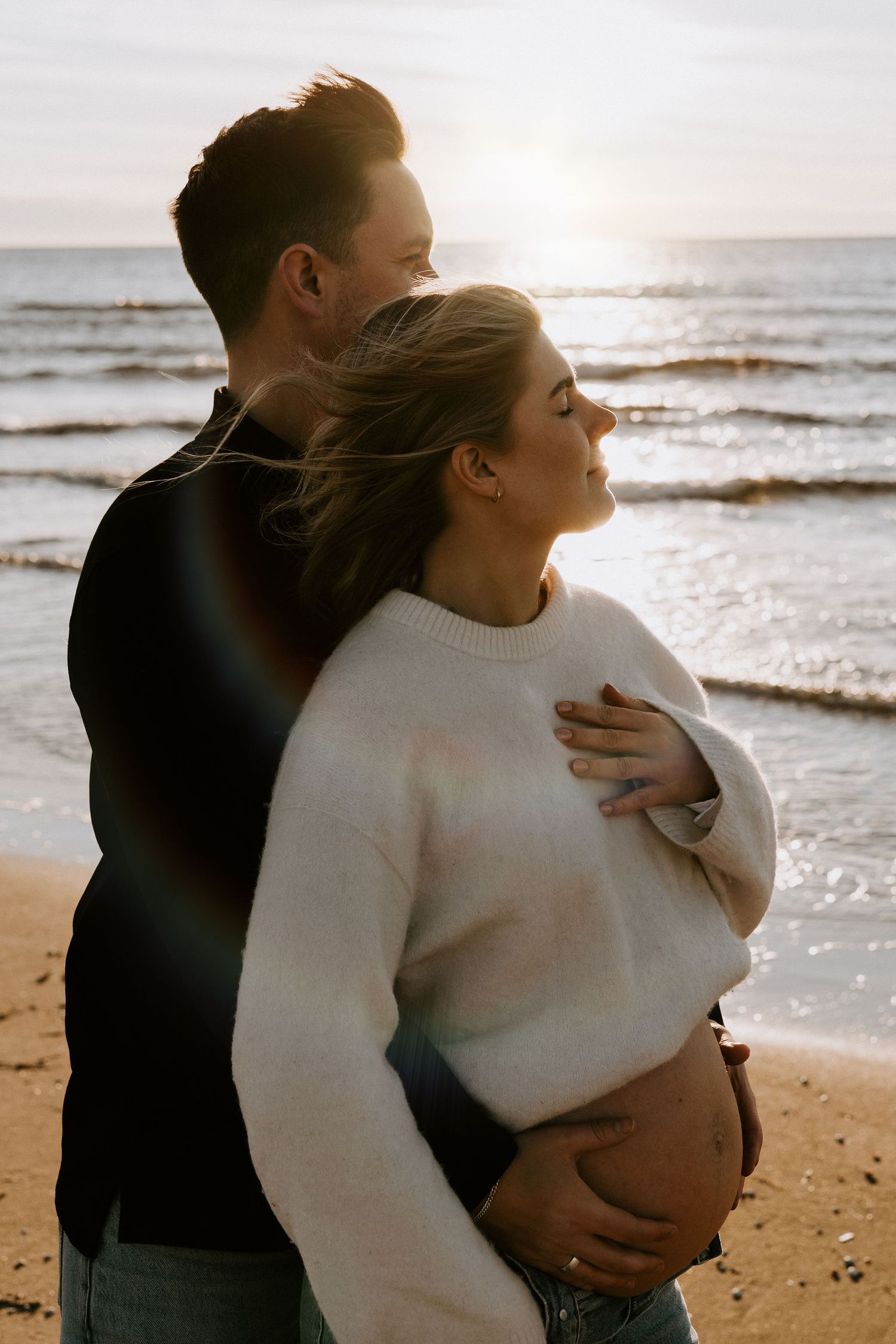 A couple embraces on a sandy beach at sunset while waves crash on the shore in the background.