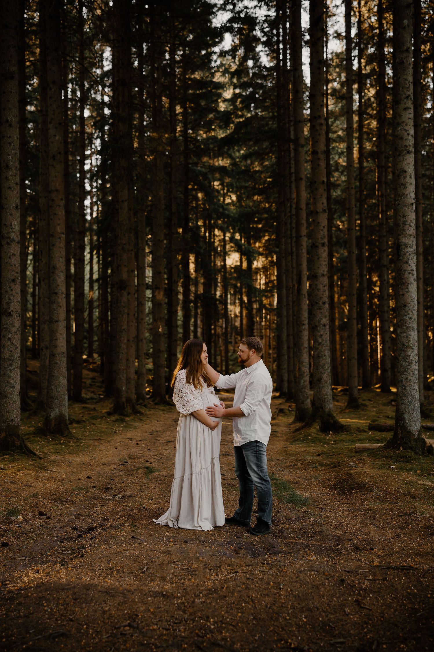 A couple shares an intimate moment in a sunlit pine forest while wearing white and casual attire.