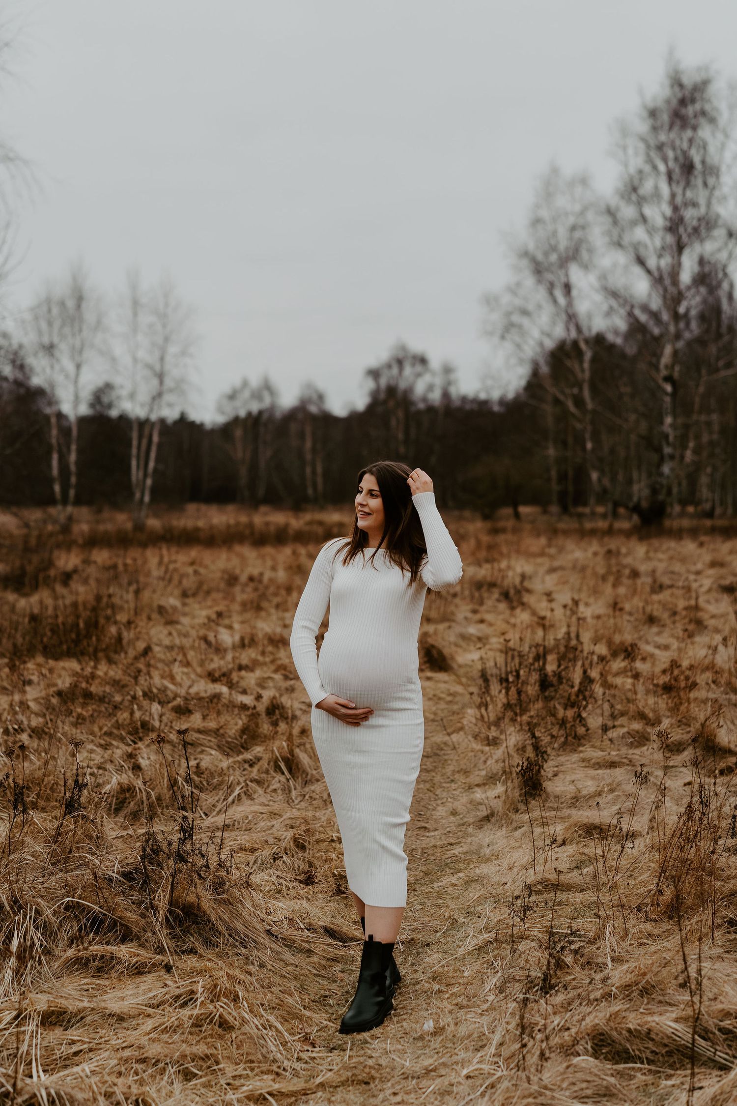 Woman in white dress poses for maternity photos in a field of dried grass with birch trees in the background.