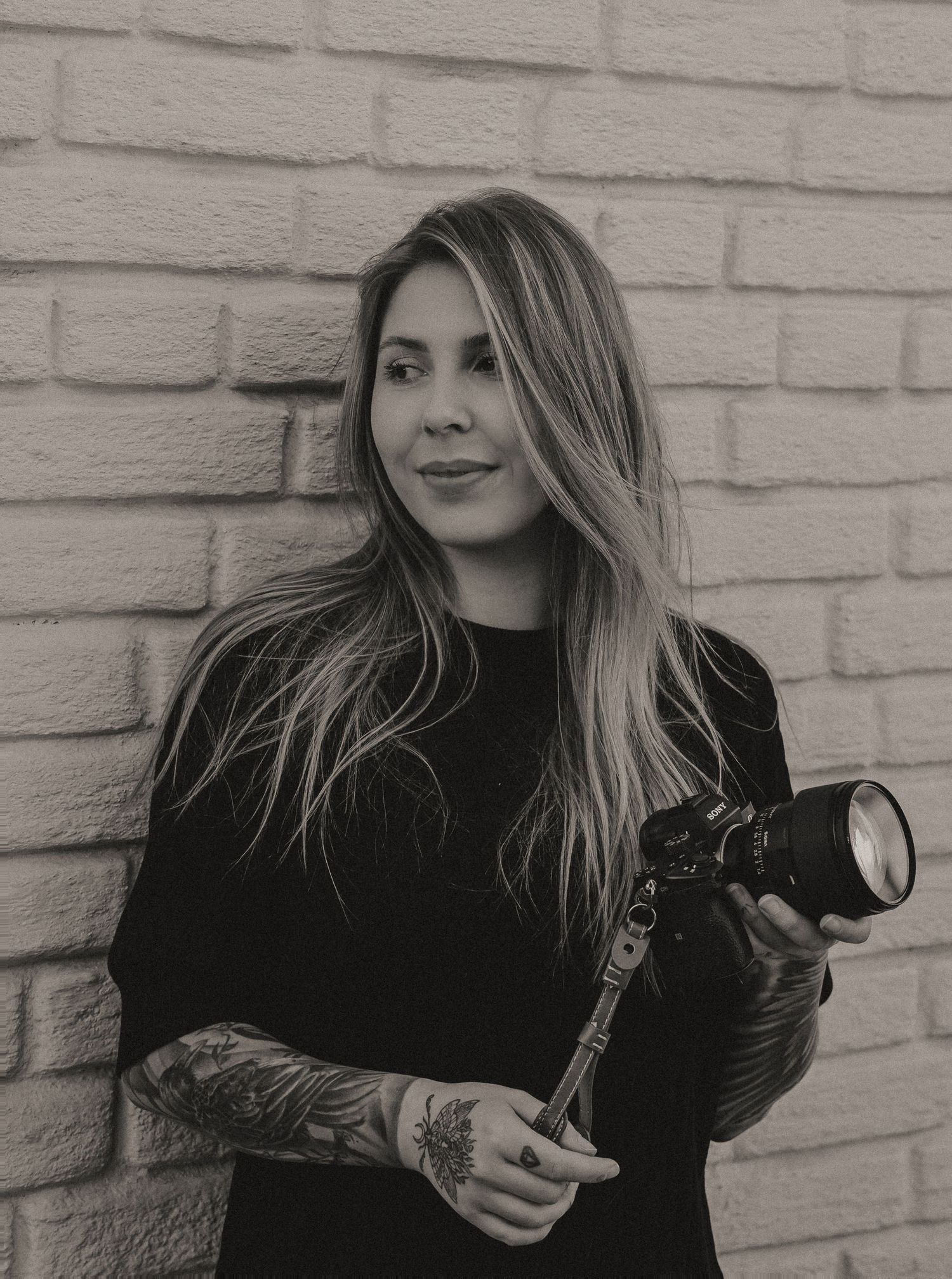 Black and white portrait of a photographer with long hair and tattoos holding a camera against a brick wall background.