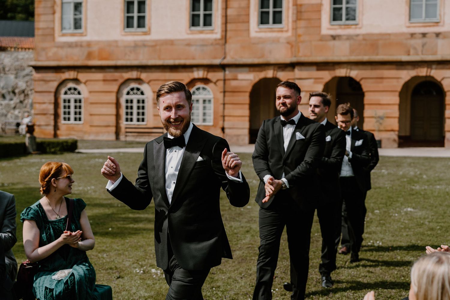 Wedding celebration scene with people in formal attire including black tuxedos on sunny day at historic building courtyard.