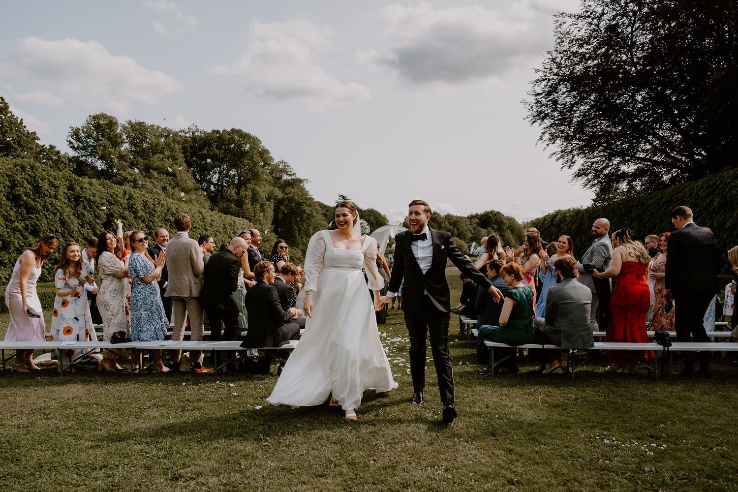 Newlyweds walking down the aisle as wedding guests stand and cheer on benches outdoors.