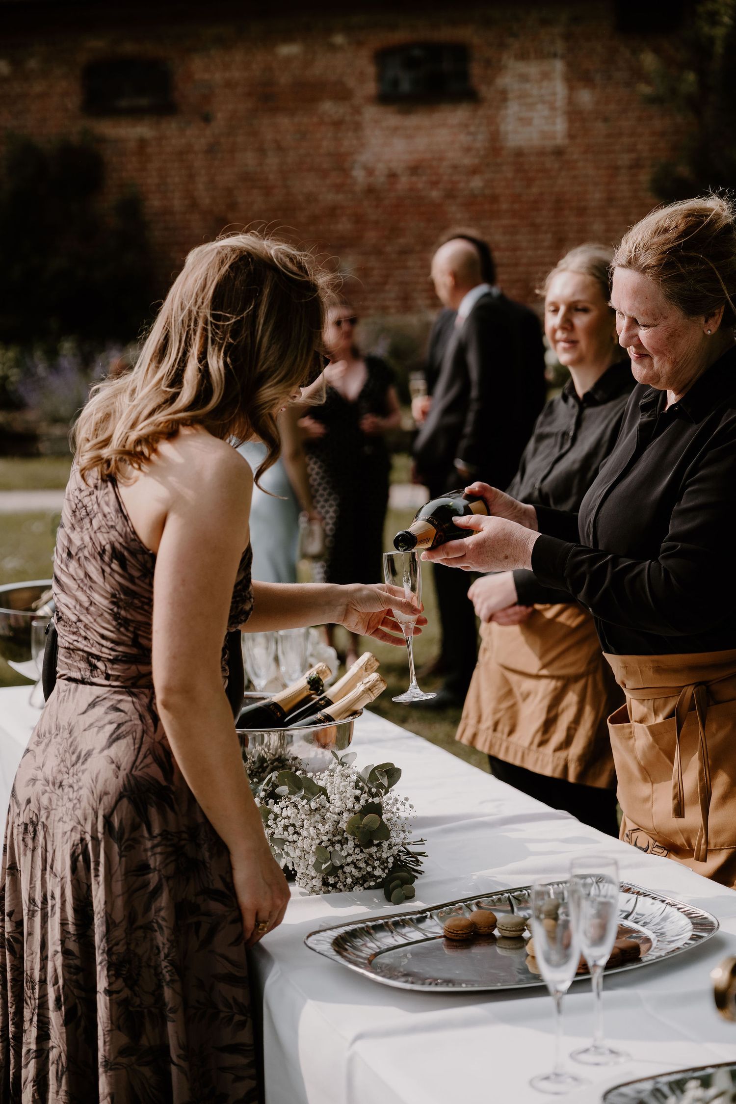 Guests and servers interact at an elegant outdoor catering event with a white tablecloth and rustic brick wall backdrop.