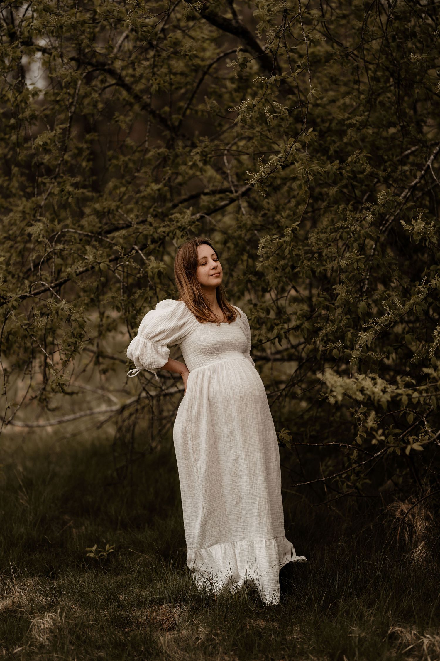 Person in a flowing white prairie dress stands gracefully among blooming spring trees in a moody nature setting.