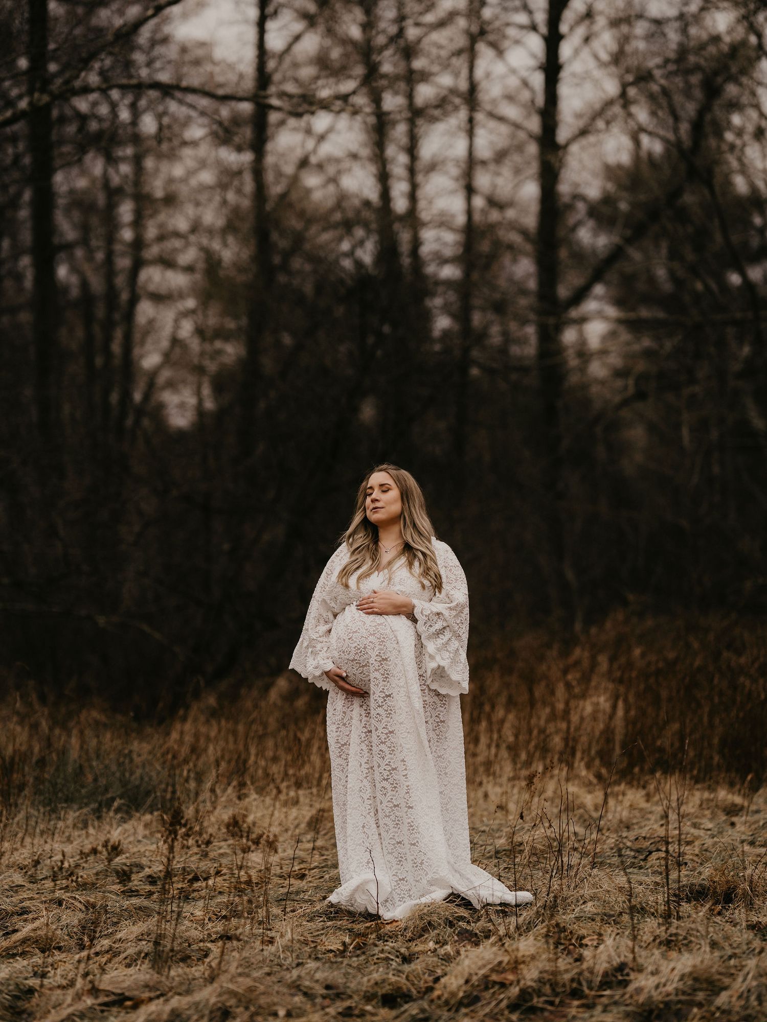A figure in a flowing white lace gown stands in a misty wooded area during golden hour.