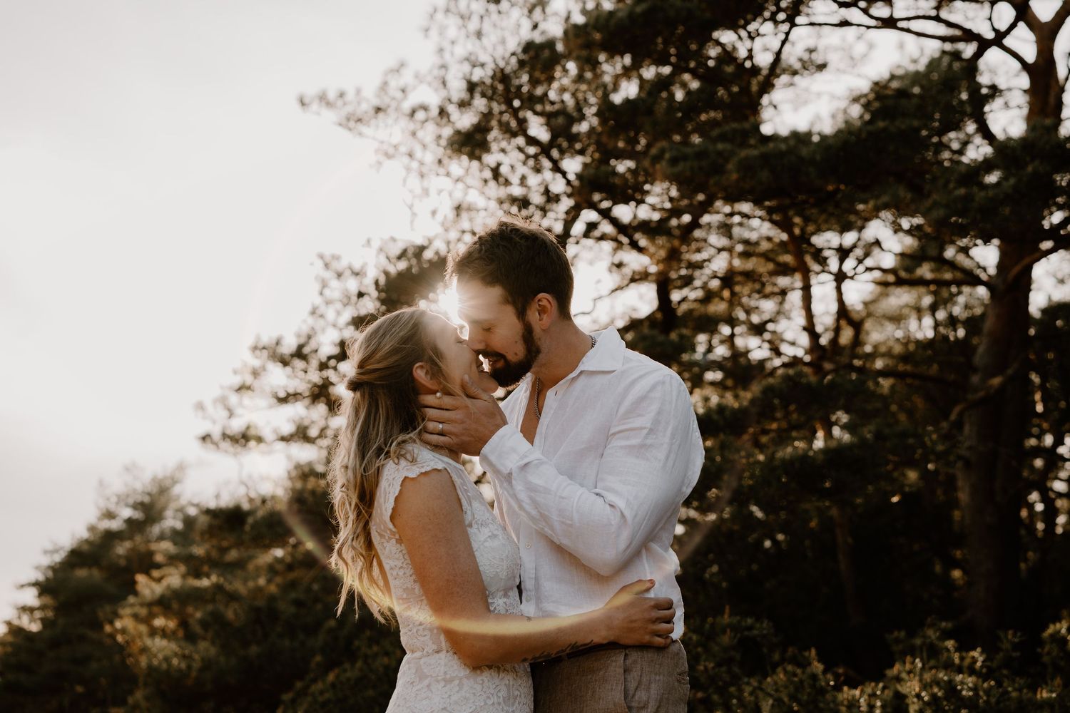 Romantic couple embraces and kisses during golden hour sunset in front of silhouetted pine trees.