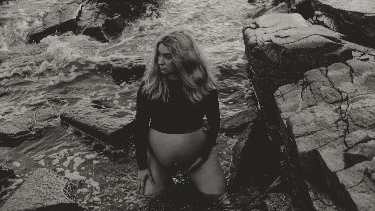 Black and white photo shows a person perched on rocky coastal terrain beside rippling ocean water.