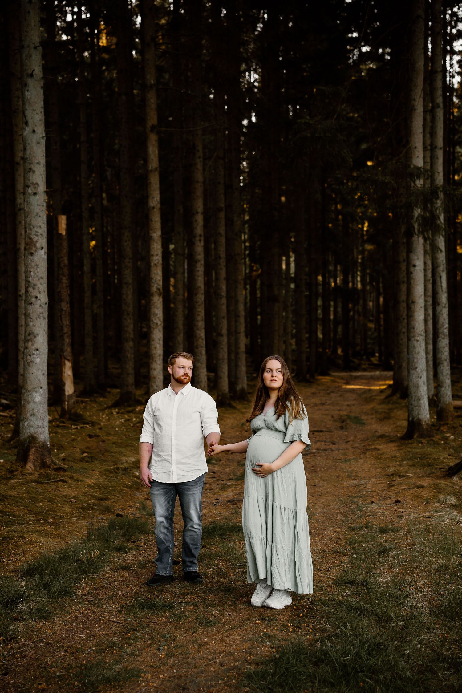 A couple stands holding hands in a towering forest with golden sunlight filtering through the trees.