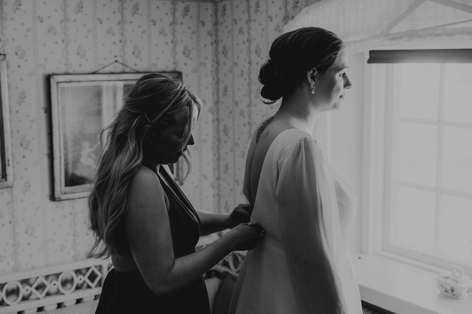 A helper assists with buttoning up a wedding dress in a softly lit black and white photograph.