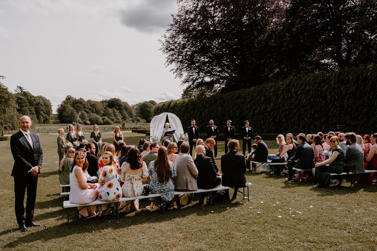 Outdoor wedding ceremony with guests sitting on rows of chairs surrounded by hedges and trees.