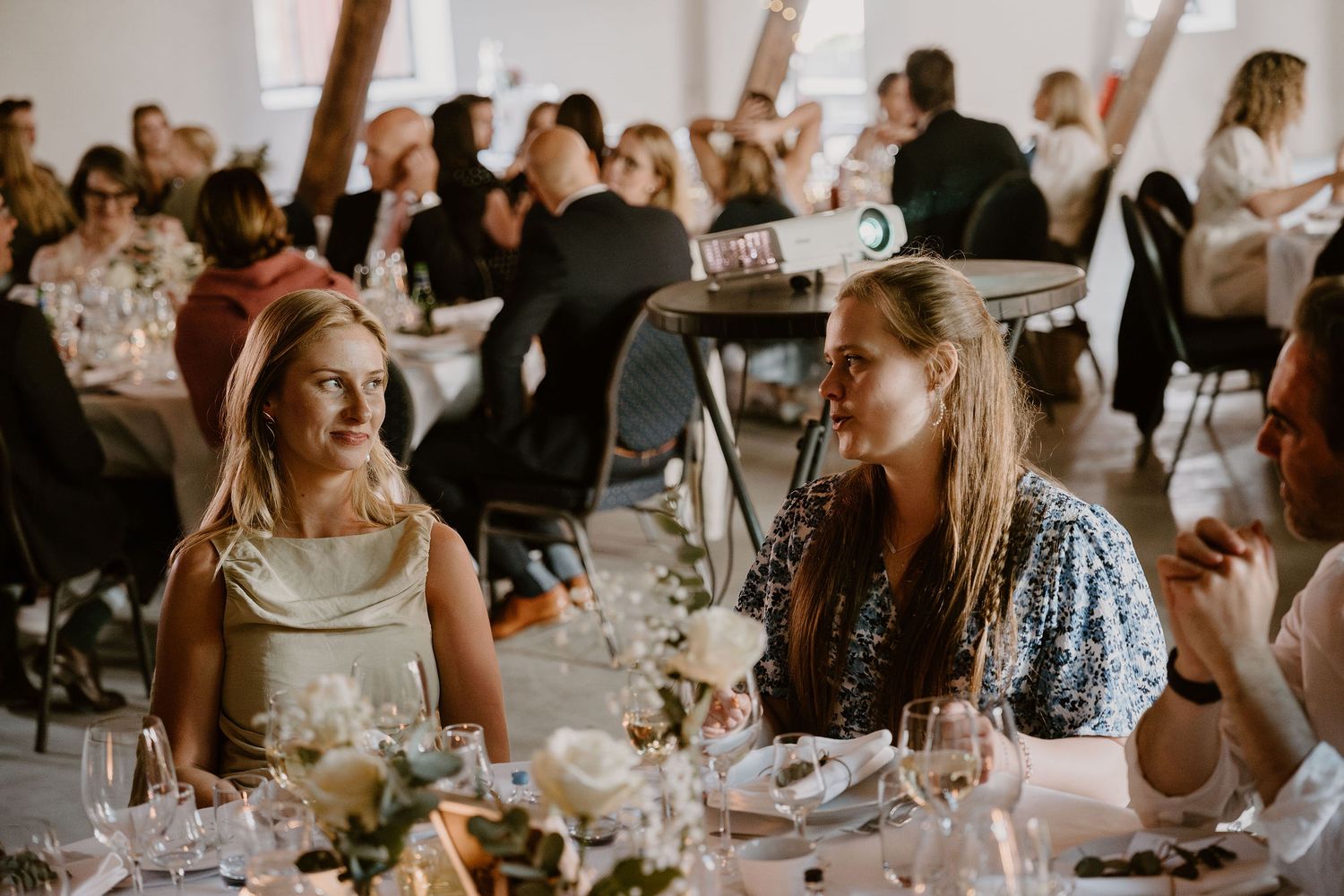 Guests mingle and converse while seated at an elegant wedding reception dinner with soft lighting and table decor.