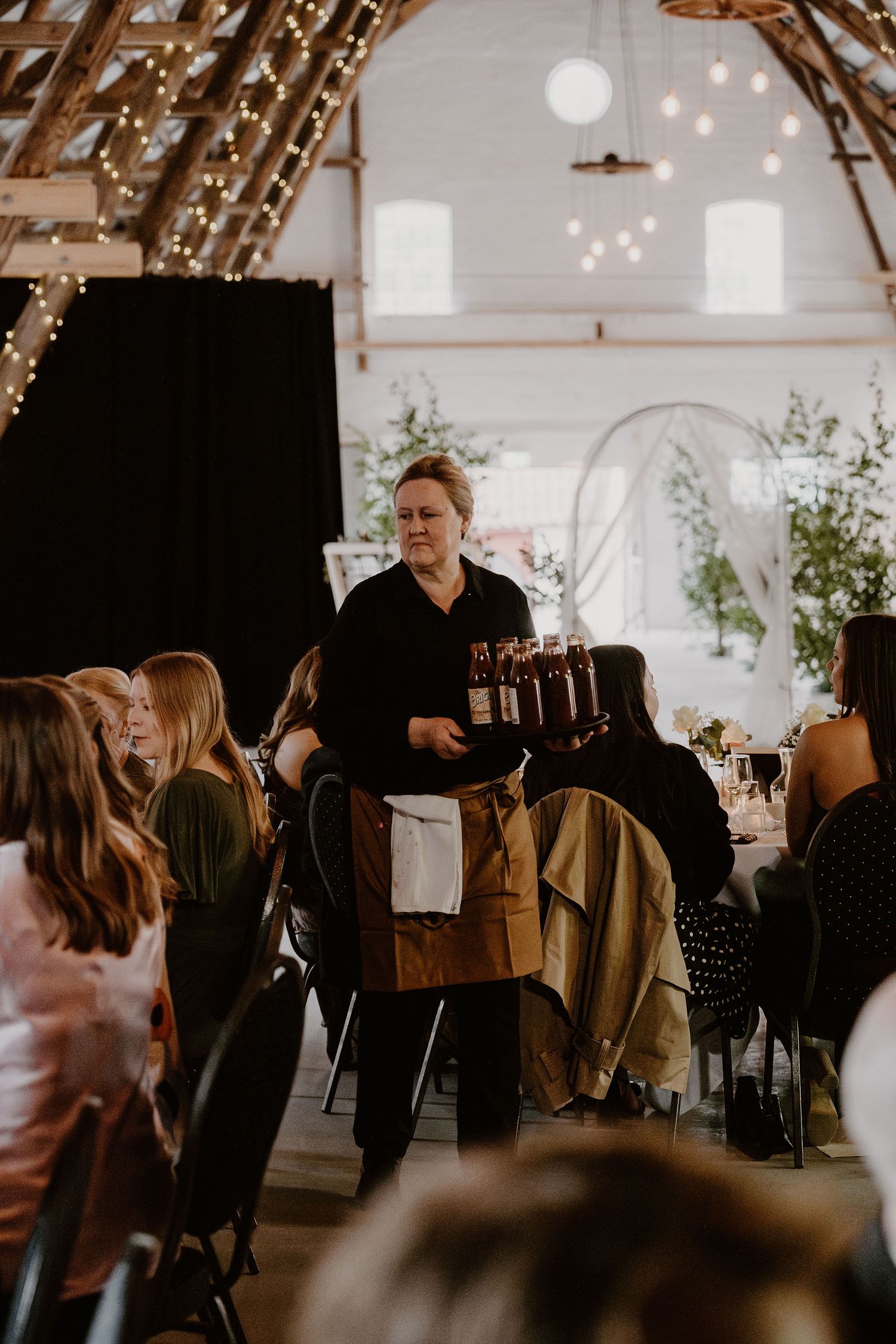 A waiter in black attire carries a large tray through an elegant reception venue decorated with string lights.