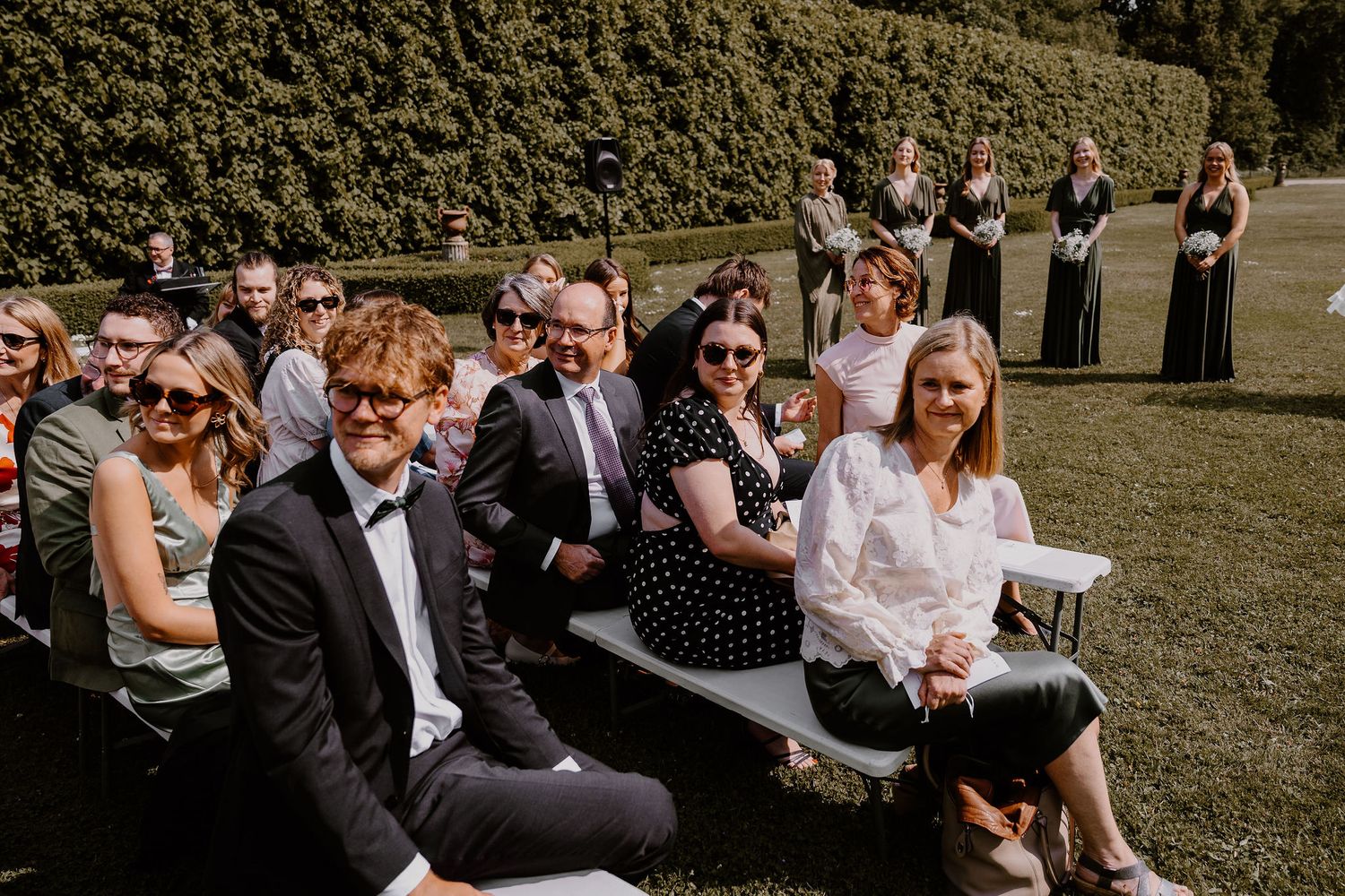 Wedding guests seated outdoors on white chairs during a sunny garden ceremony.