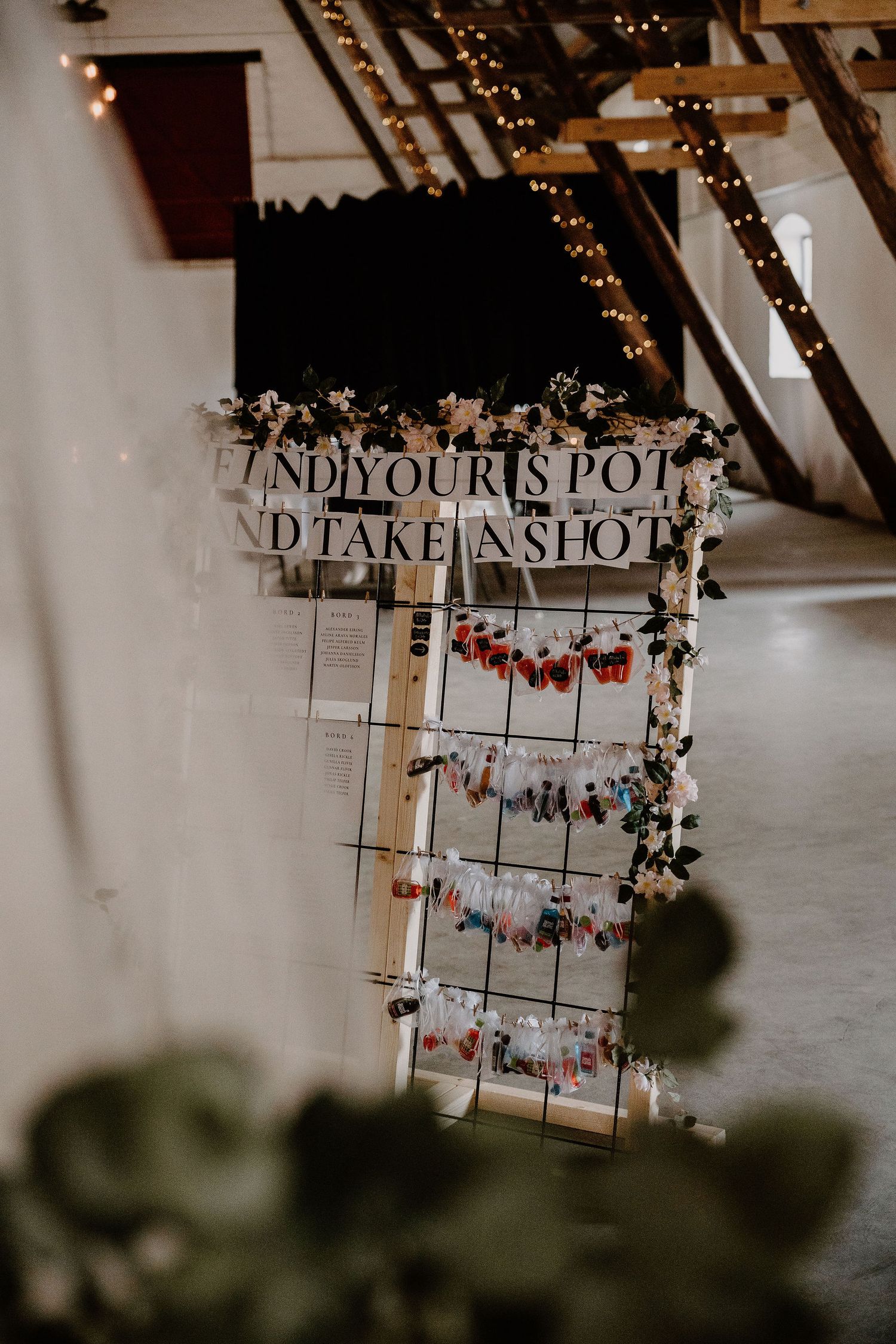 A rustic photo display grid with 'Find Your Seat' signage decorated with fairy lights and floral garlands at a wedding.