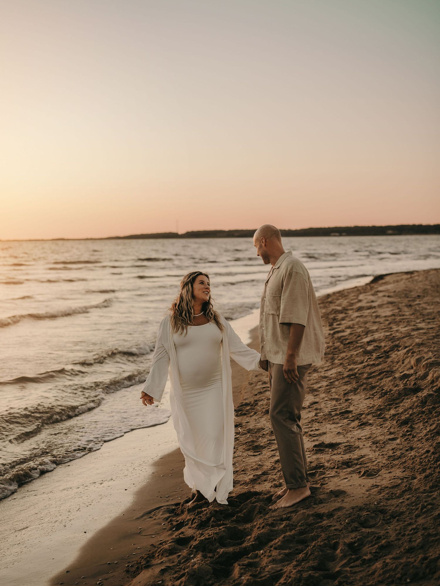 A romantic couple walks along a sandy beach at sunset, with waves lapping the shore and warm golden light bathing the scene.