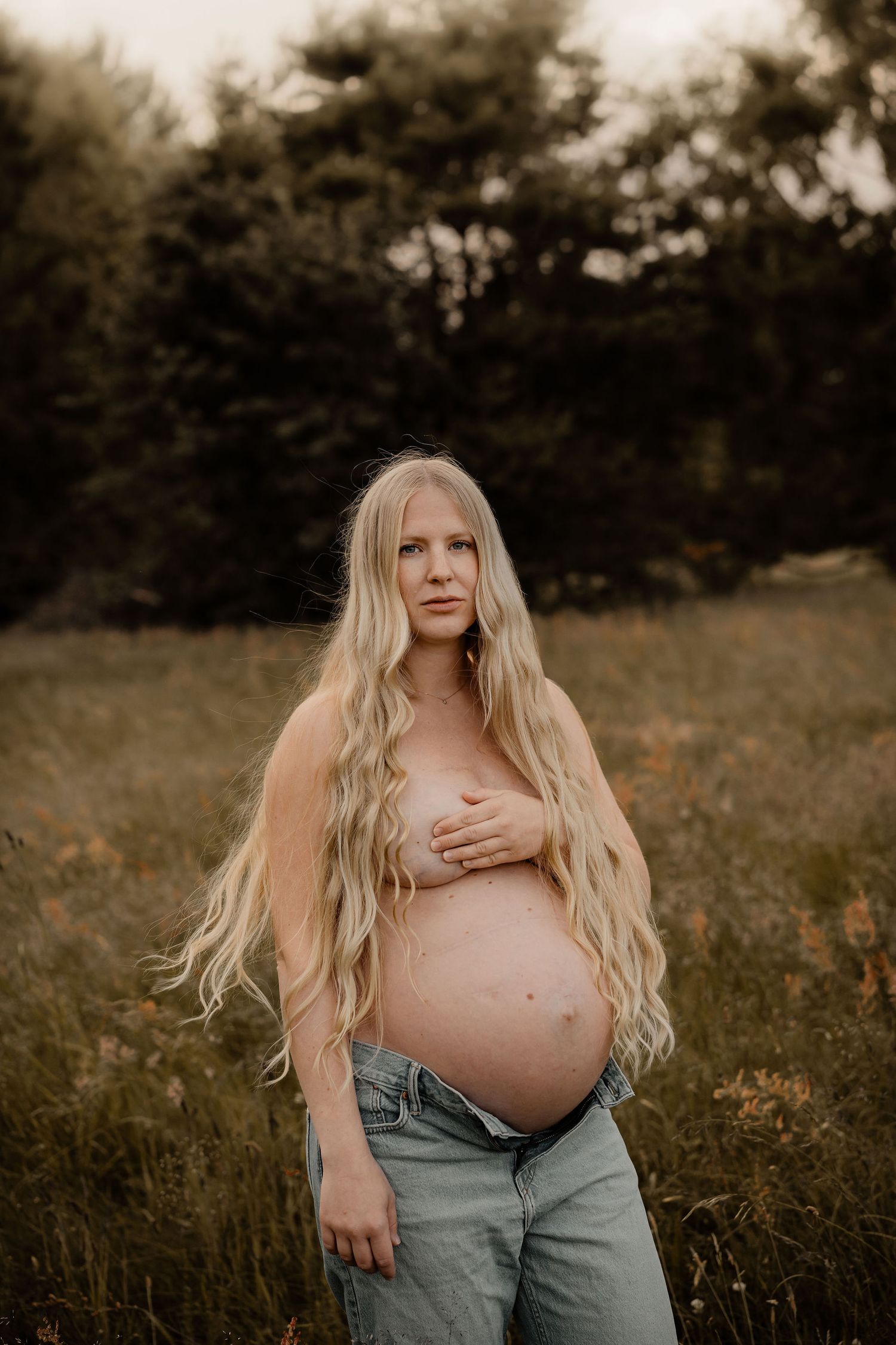 Artistic maternity photo of an expectant mother with long blonde hair standing in a sunlit meadow at golden hour.