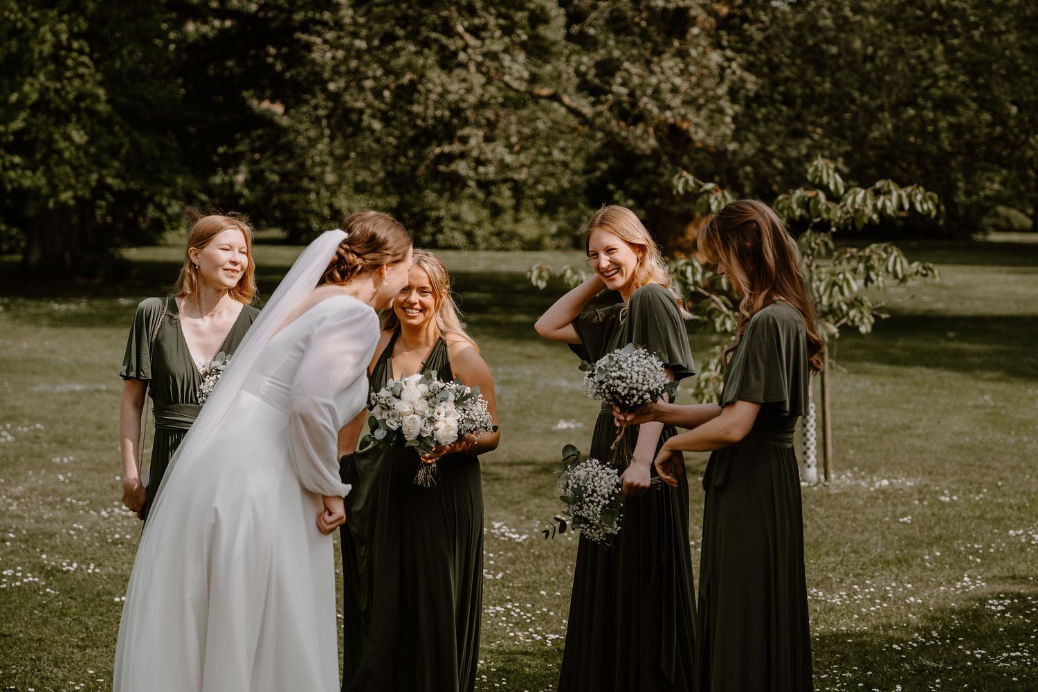 A bride and bridesmaids in dark dresses share a candid laughing moment during an outdoor wedding photoshoot.
