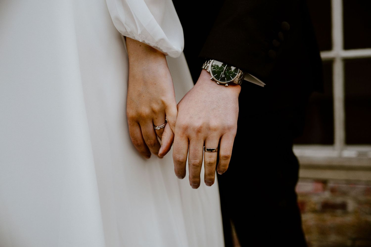 Close-up shot of couple's hands with wedding rings, white dress sleeve and black suit visible.