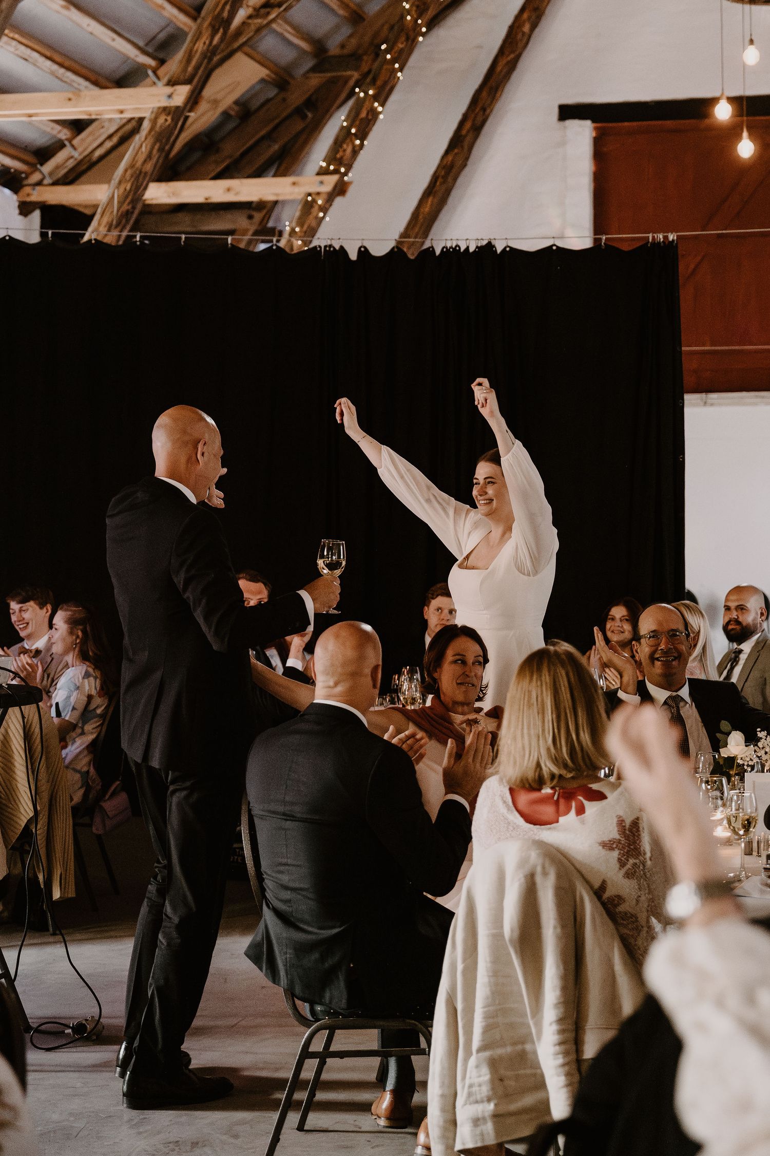 Wedding reception celebration with guests raising glasses during a toast in rustic venue with exposed wooden beams.
