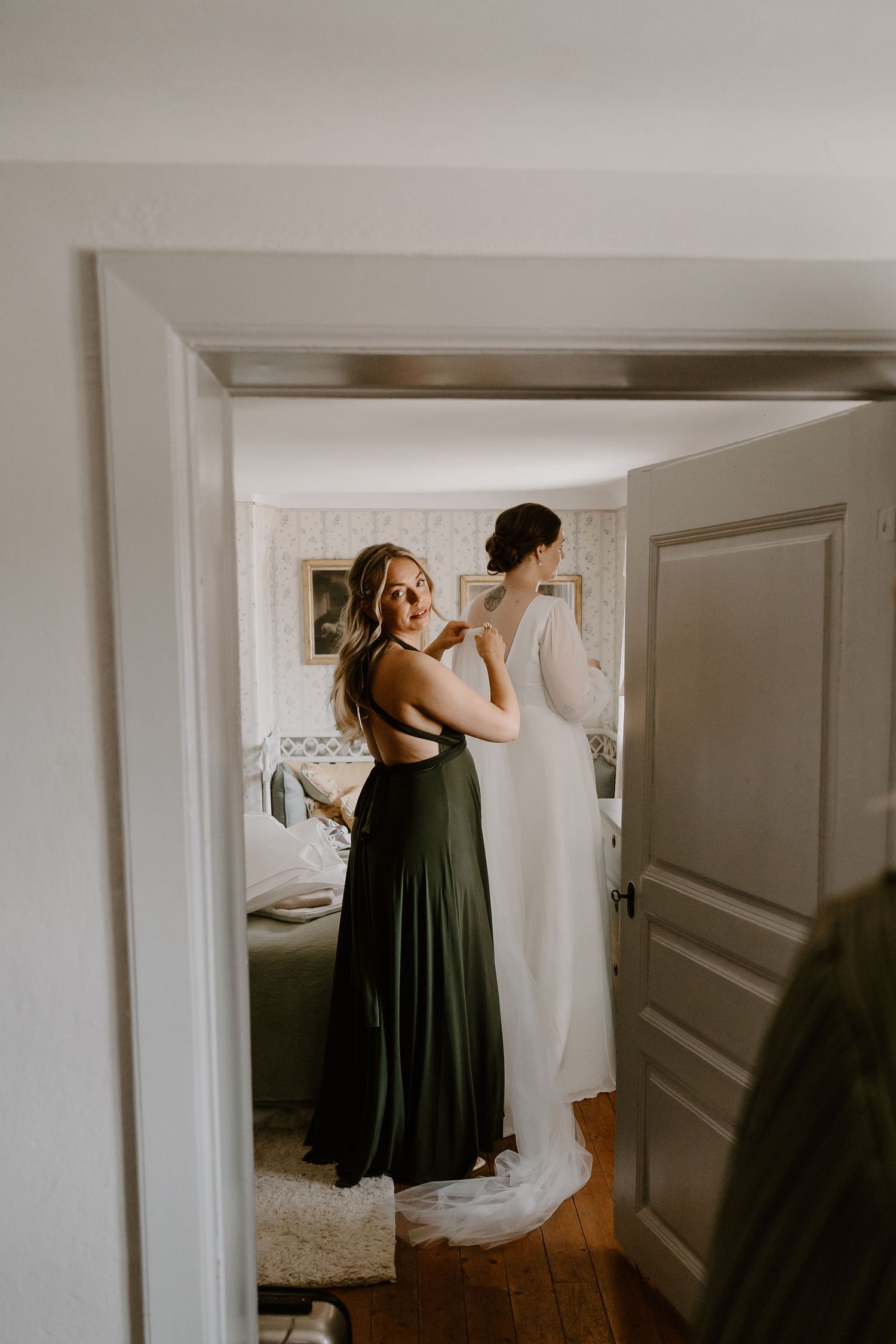 Person wearing a flowing dark dress looks at two white wedding dresses hanging in a softly lit dressing room.