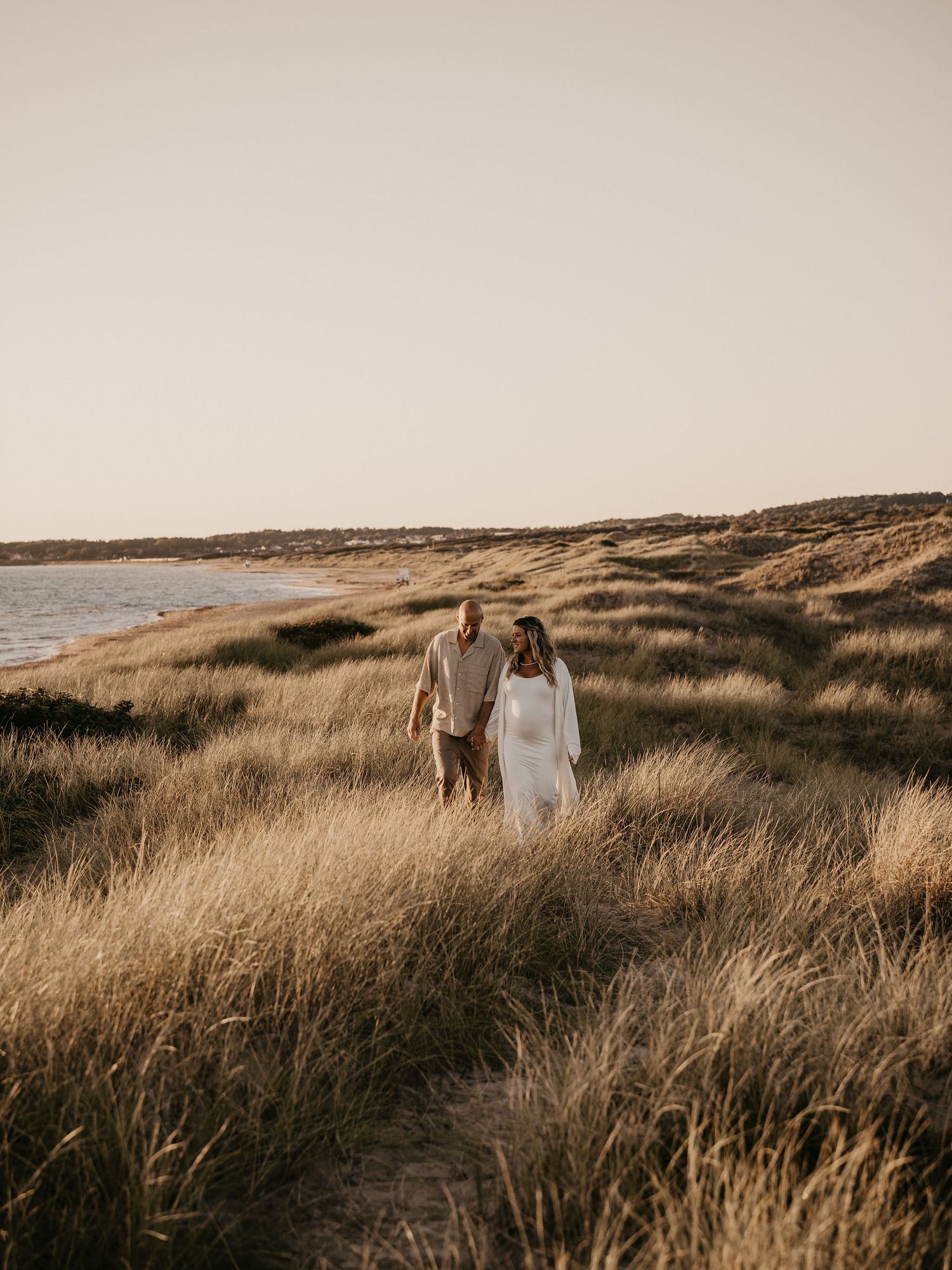 A couple stands together in golden coastal grass at sunset along a scenic beach shoreline.