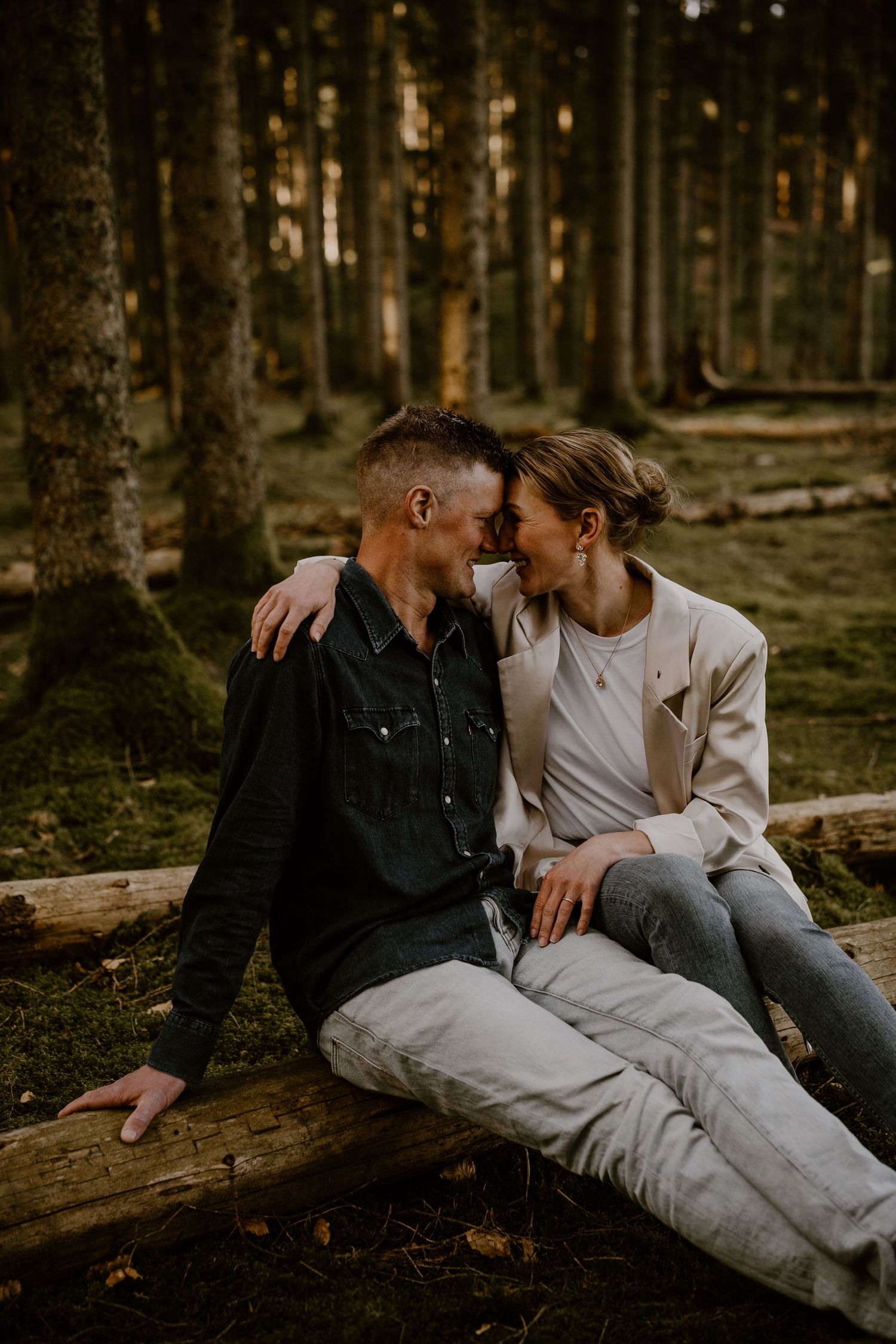 A loving couple shares an intimate moment while sitting on a fallen log in a moody forest setting at sunset.