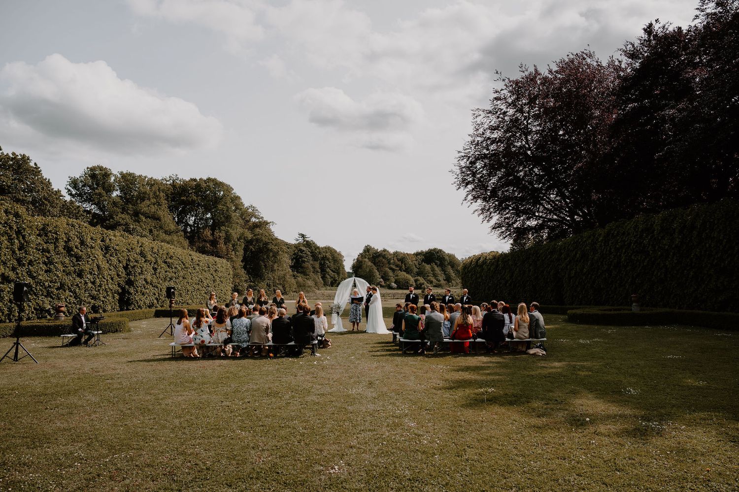 An outdoor wedding ceremony in a scenic garden setting under cloudy skies with guests seated on either side of the aisle.