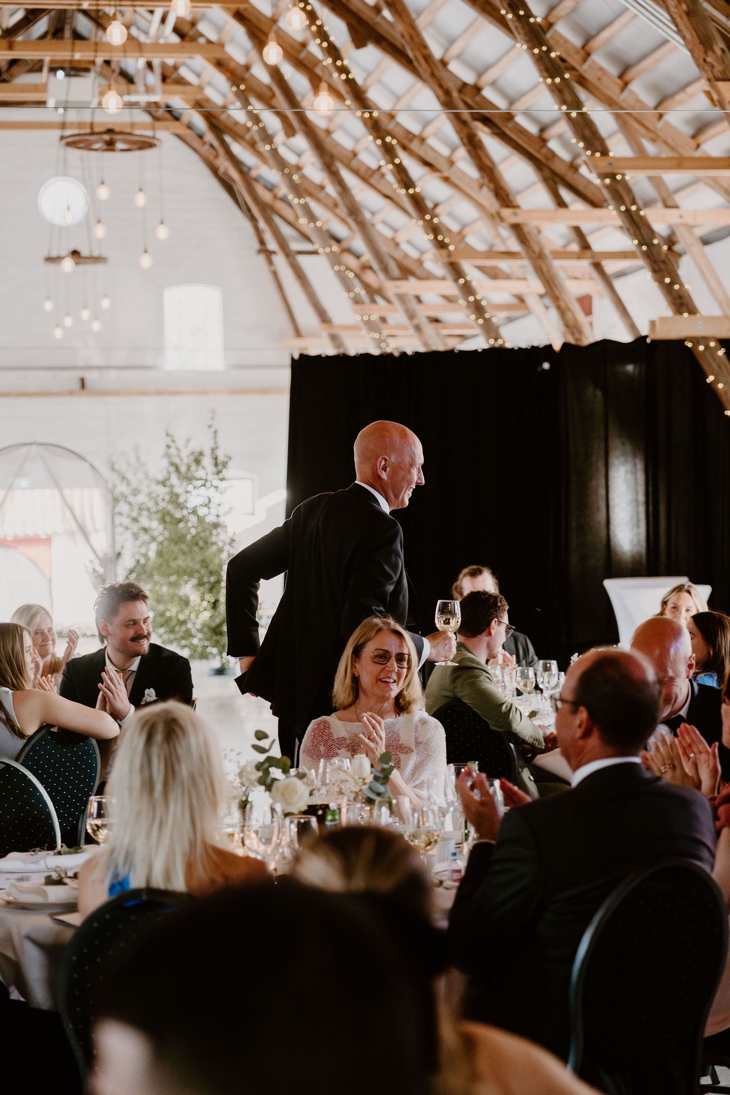 A wedding reception in a rustic venue with exposed wooden beams and string lights decorating the vaulted ceiling.