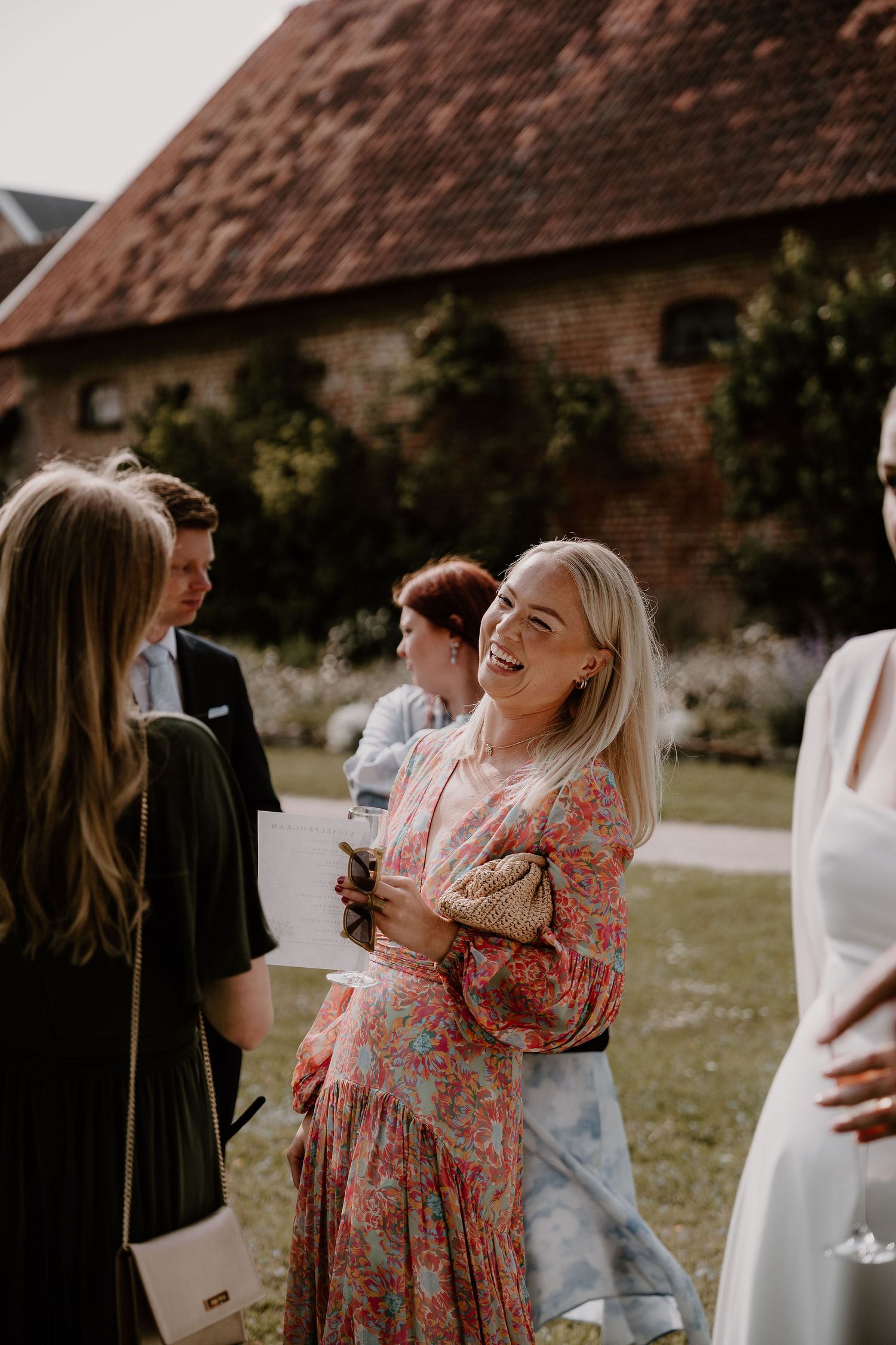 A group in a floral dress and dark attire socializing at an outdoor gathering near a rustic brick building.