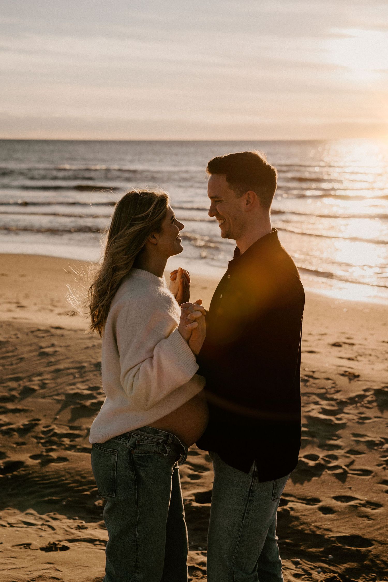 A couple shares an intimate moment on the beach at sunset, standing close together in casual sweaters and jeans.