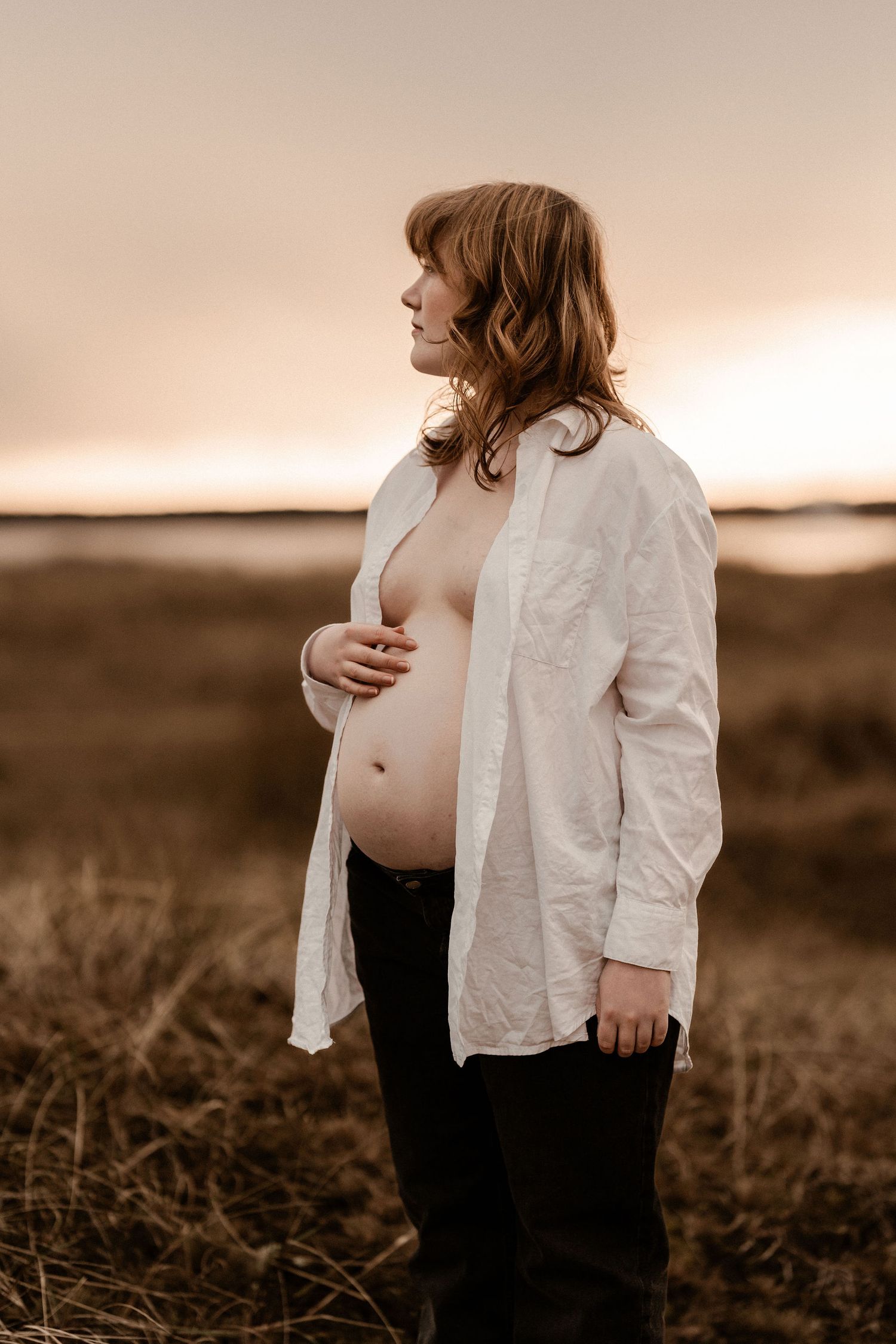 A pregnant woman in a white button-down shirt stands in a field against a sunset sky during a maternity photoshoot.
