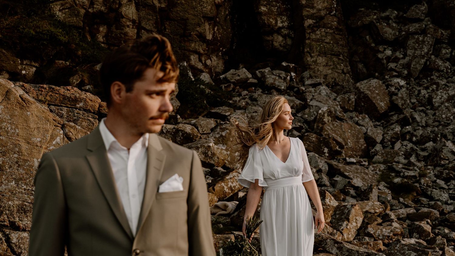 A couple in wedding attire stands against a rocky cliff backdrop during a golden hour photoshoot.