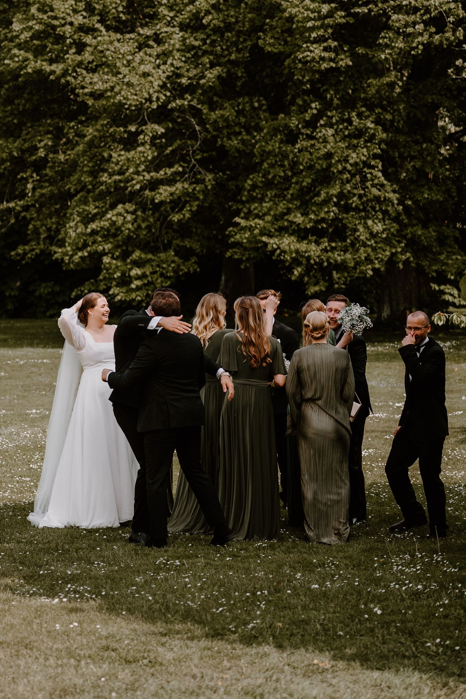 Wedding party poses together for fun photoshoot in a field with trees in the background, showing joyful and playful moments.