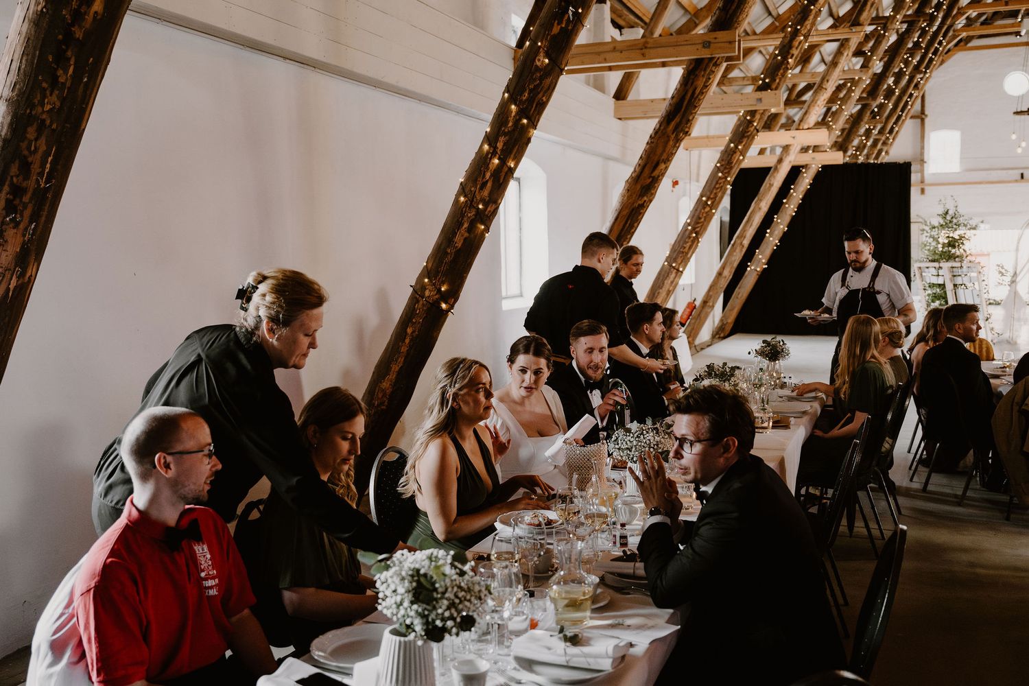 Wedding guests enjoying cocktail hour and mingling at a rustic indoor reception venue with dark wooden beams.