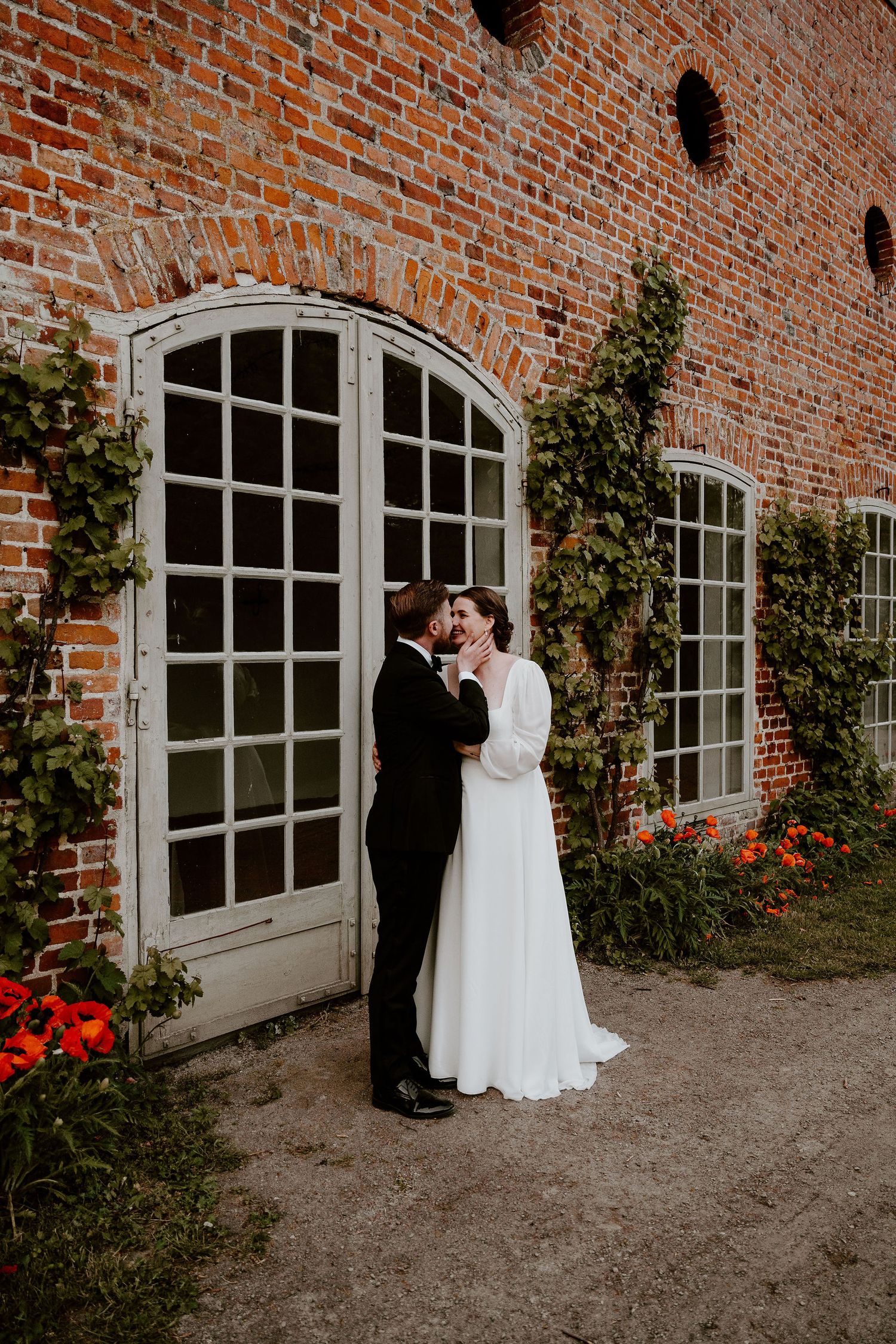 A couple shares intimate moments against a rustic brick building with arched windows and climbing vines in a romantic setting.