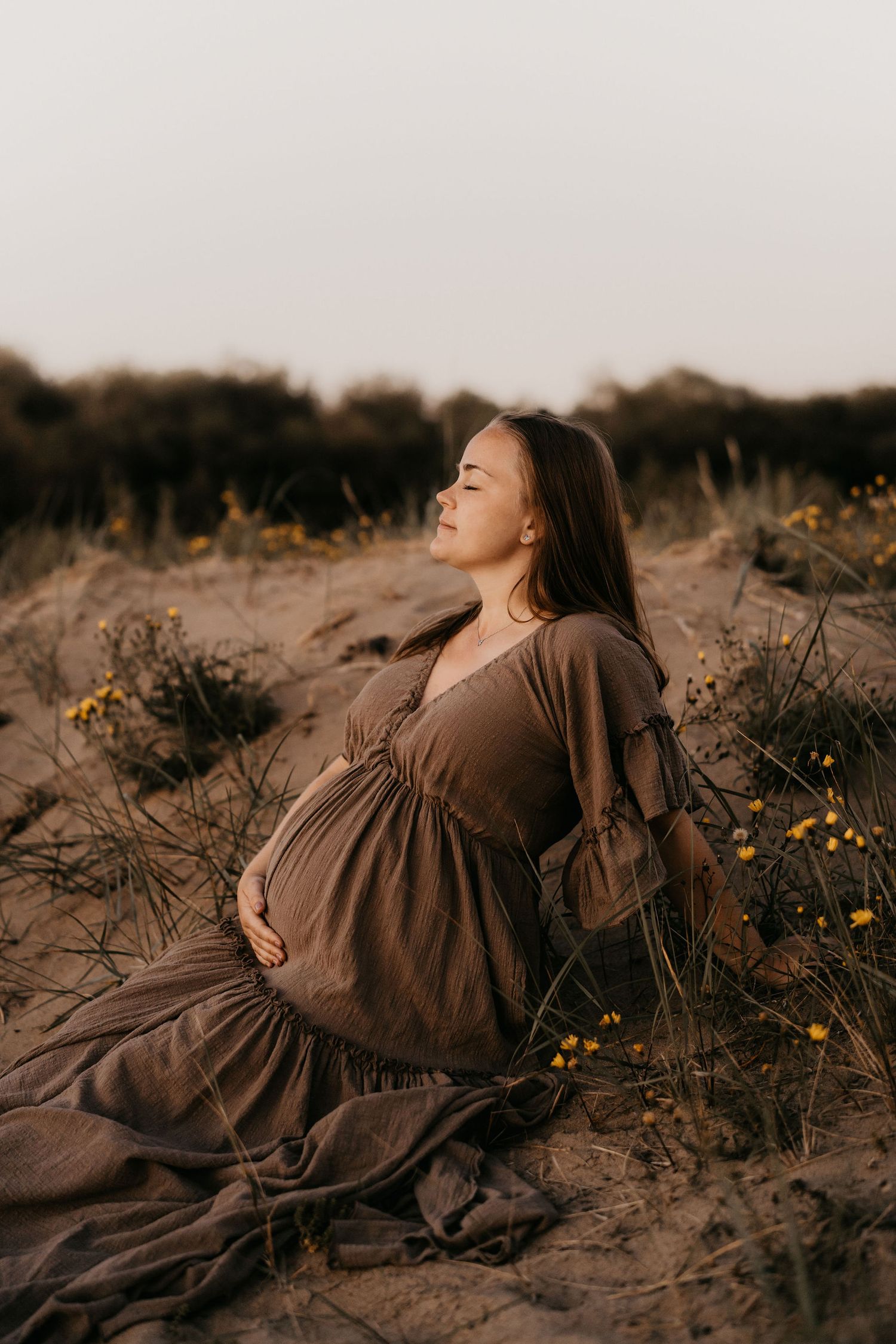 Expectant mother in flowing brown dress sits peacefully among wildflowers during golden hour sunset maternity photoshoot.